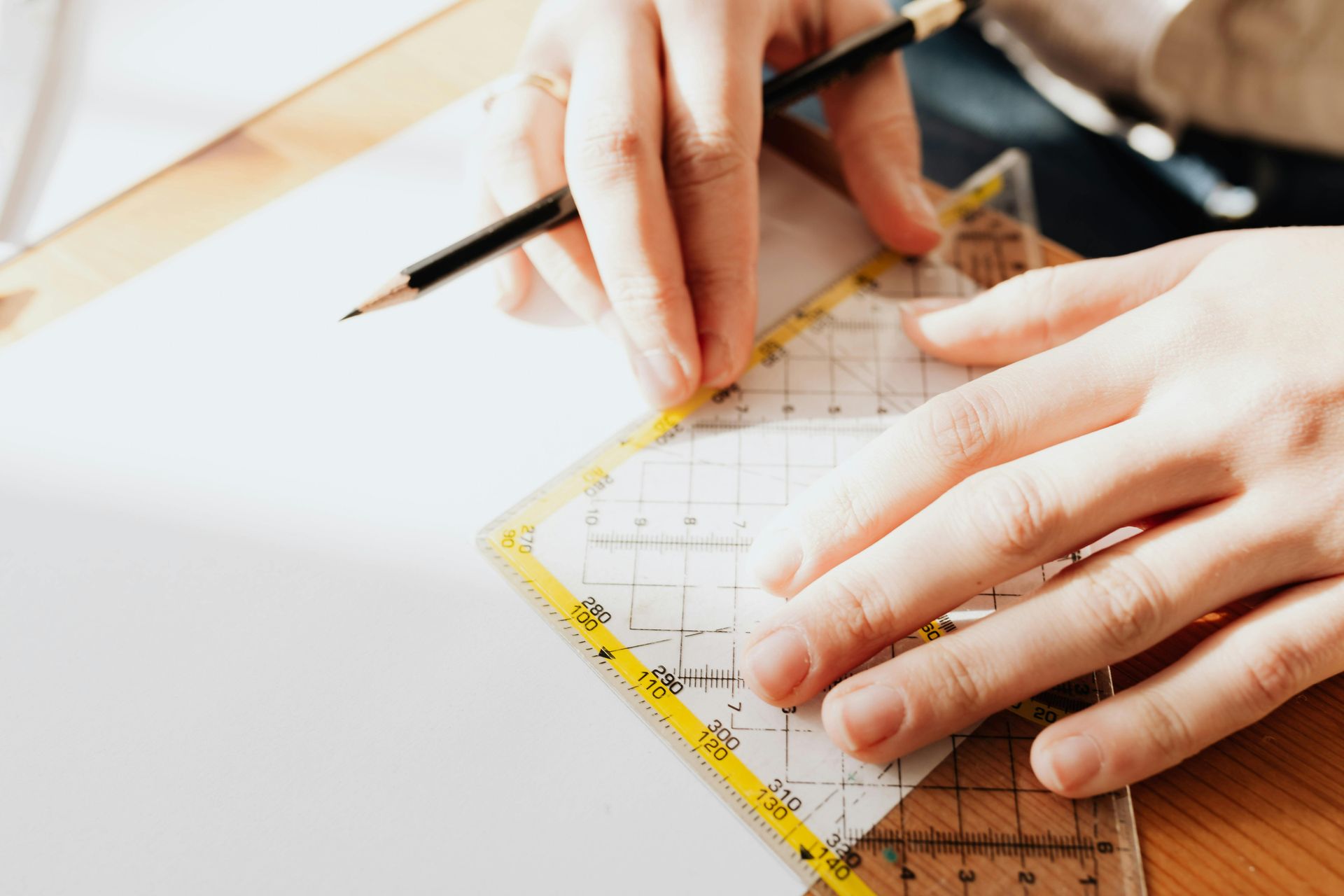 A pair of hands uses a black pencil and a clear, yellow-bordered grid ruler to draw lines on a white sheet of paper.