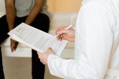 A person in a white lab coat holds a clipboard and pen while interacting with another person in a clinical setting.