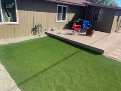 A patch of artificial grass adjacent to a tan patio with two folding chairs and a fire pit in front of a brown house.
