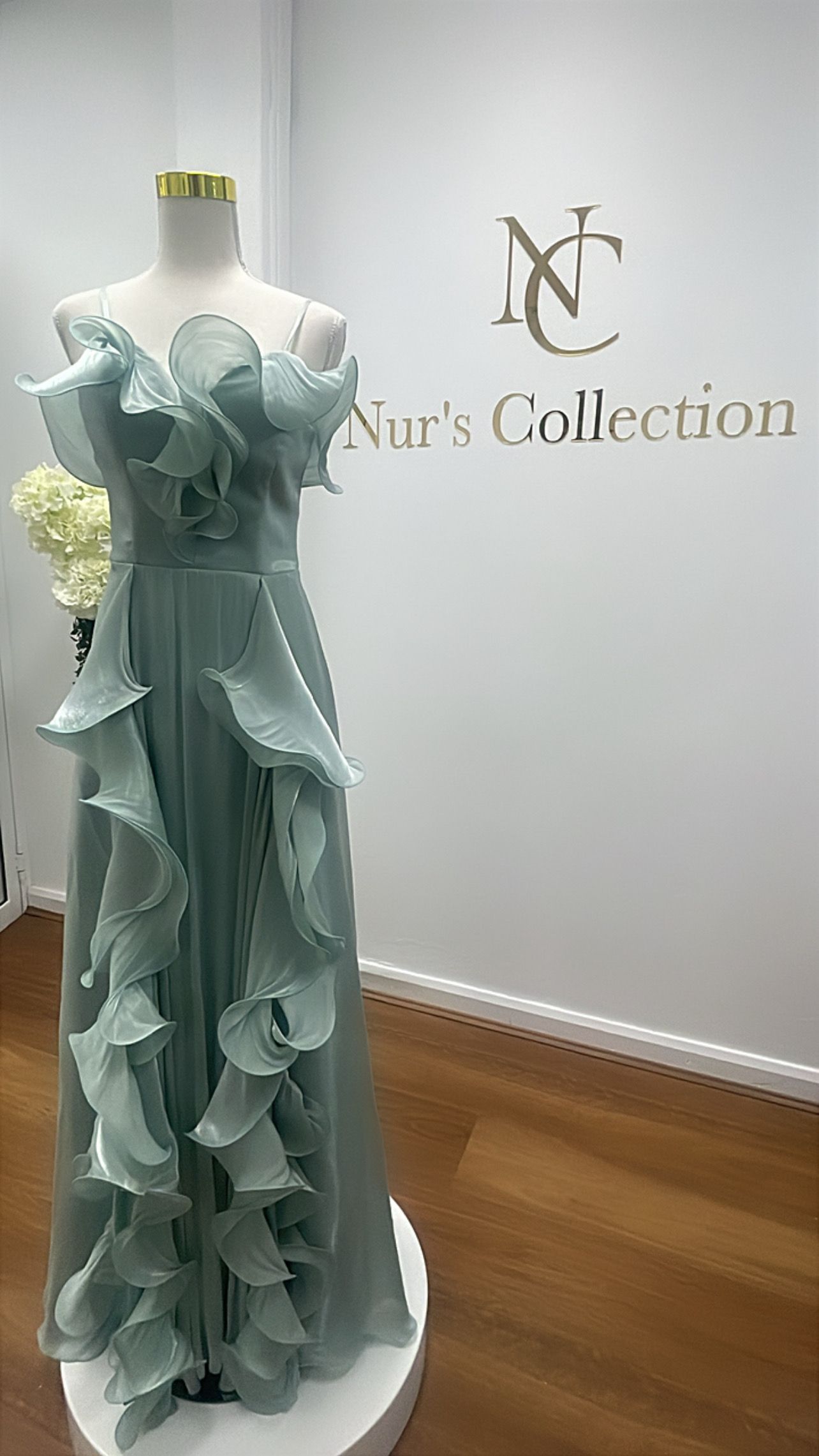 Elegant seafoam-green gown on a mannequin, featuring ruffles. Beside a wall with a logo: