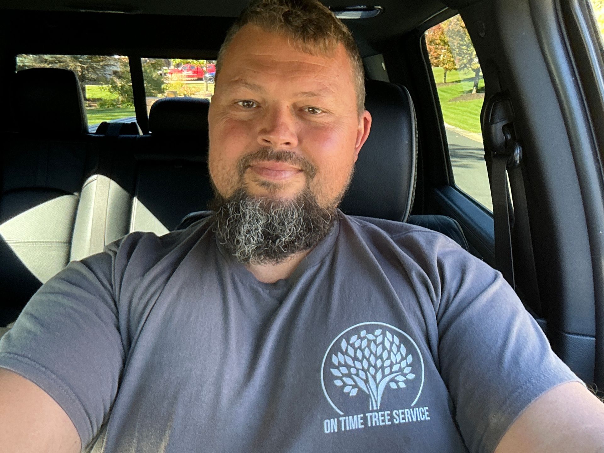 Man in gray shirt with beard sitting in a car, smiling. On Time Tree logo on shirt.