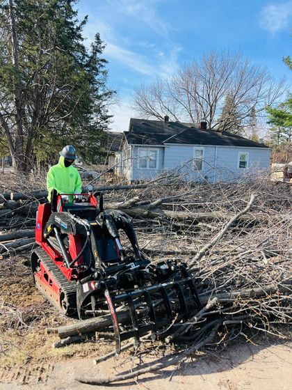 A person in a bright yellow jacket operates a red skid steer with a grapple attachment to clear fallen tree branches.