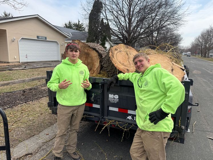 Two people in neon green hoodies stand beside a trailer filled with large logs on a residential street.