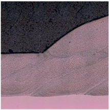 Microscopic view of a weld. Two metal layers joined; dark top layer meets a lighter layer, showing the weld's cross-section.