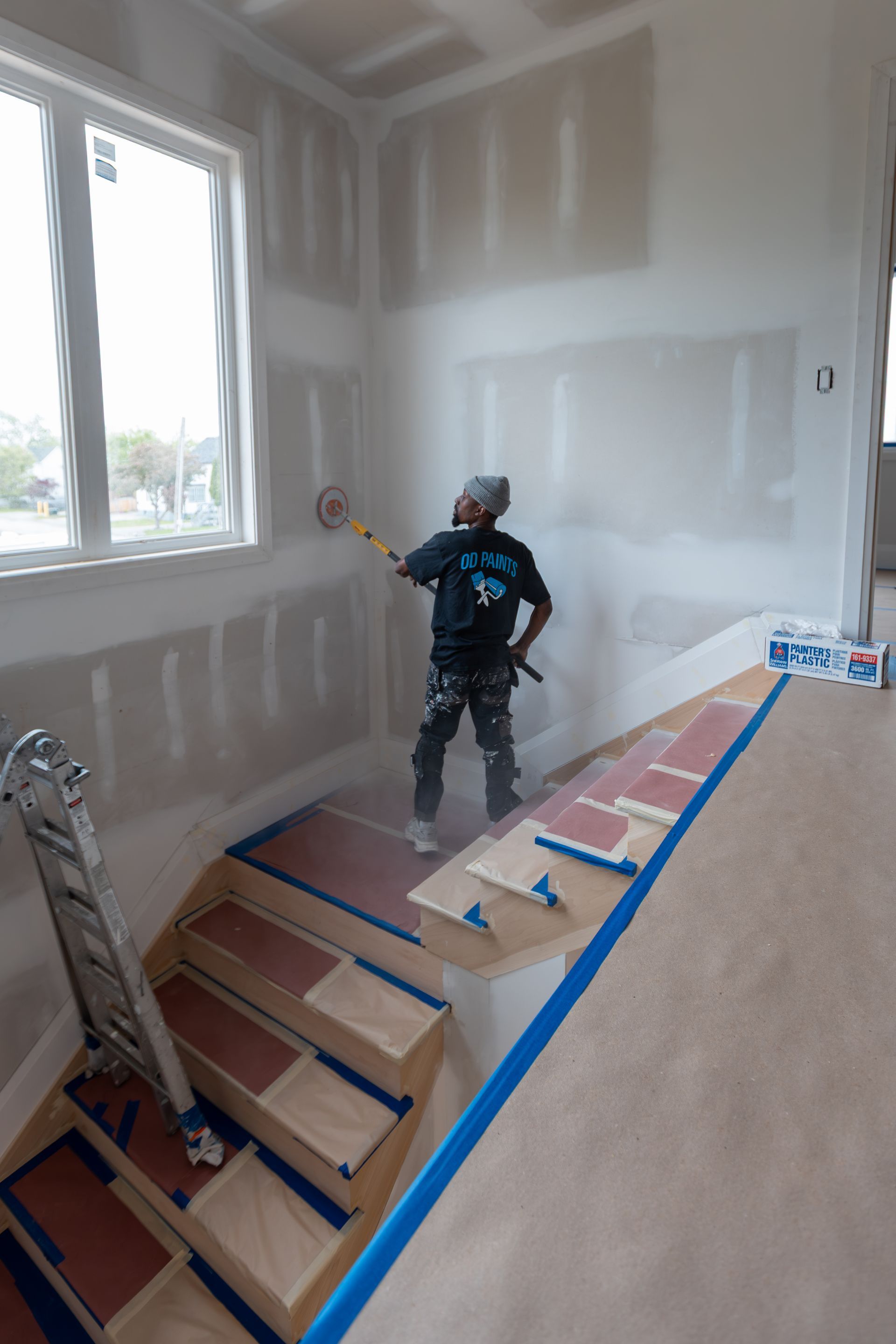 Person sanding drywall on a staircase. Interior view of new construction, with blue tape, ladder, and paint cans.