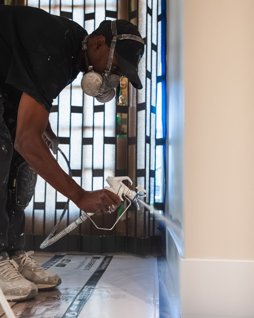 Painter spraying a wall with paint. He's wearing a respirator mask, hat, and spraying near a doorway.