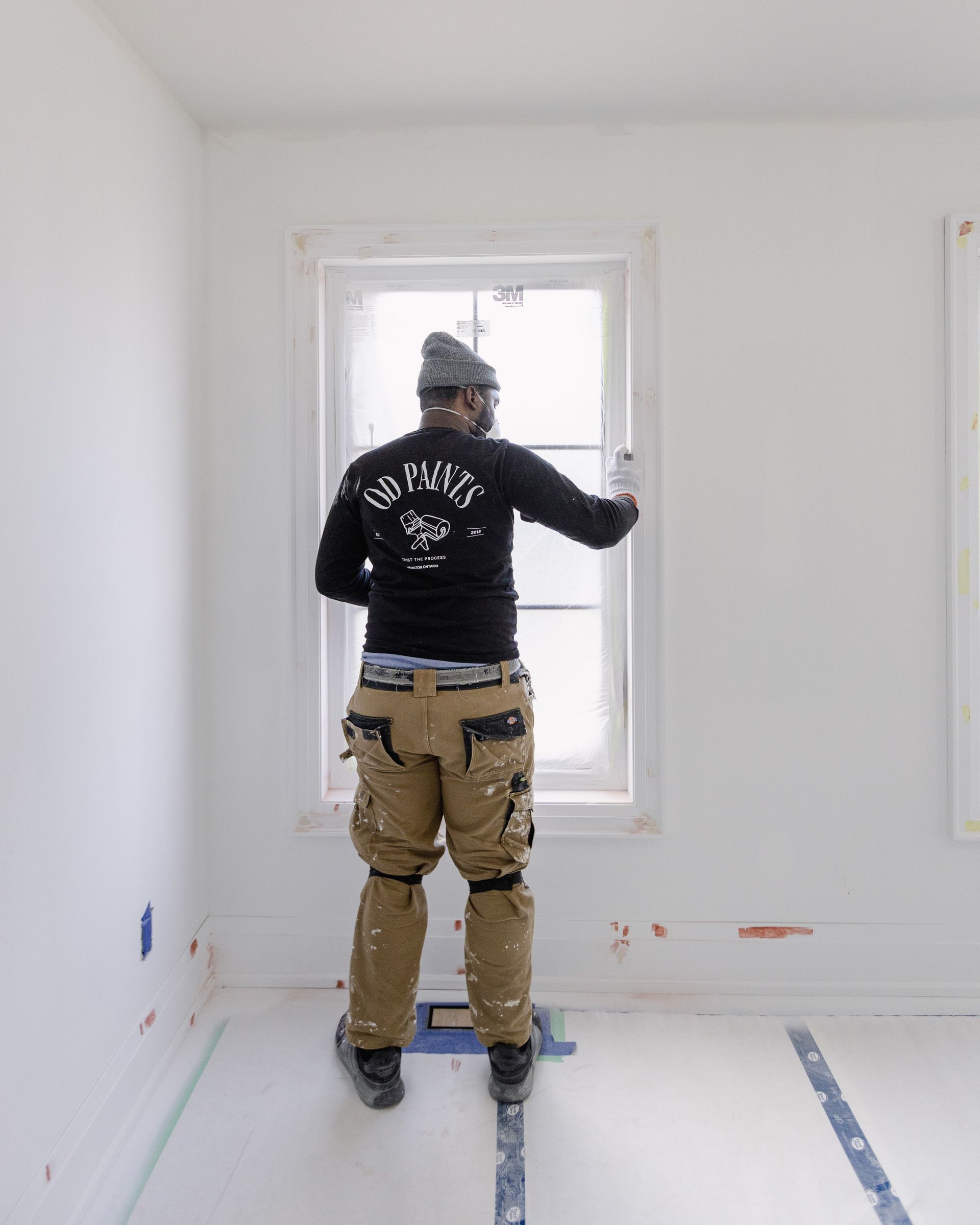 A person in work clothes inspects a window frame in a white-walled room, wearing a beanie.