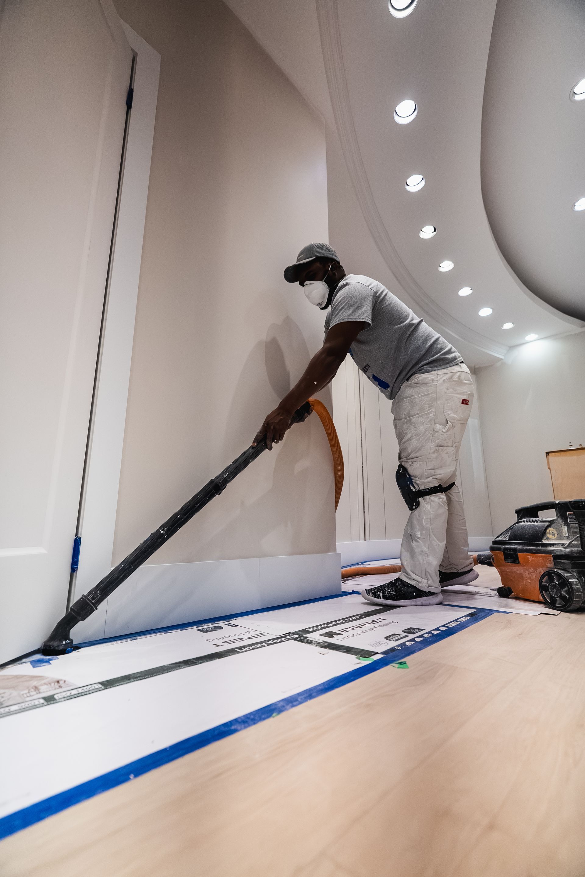 Person sanding a wall, wearing a mask and knee pads. Light room, wood floor.