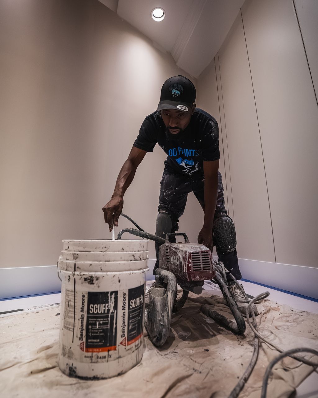 Person in black shirt using a paint sprayer in a room with white walls, beside a paint bucket.