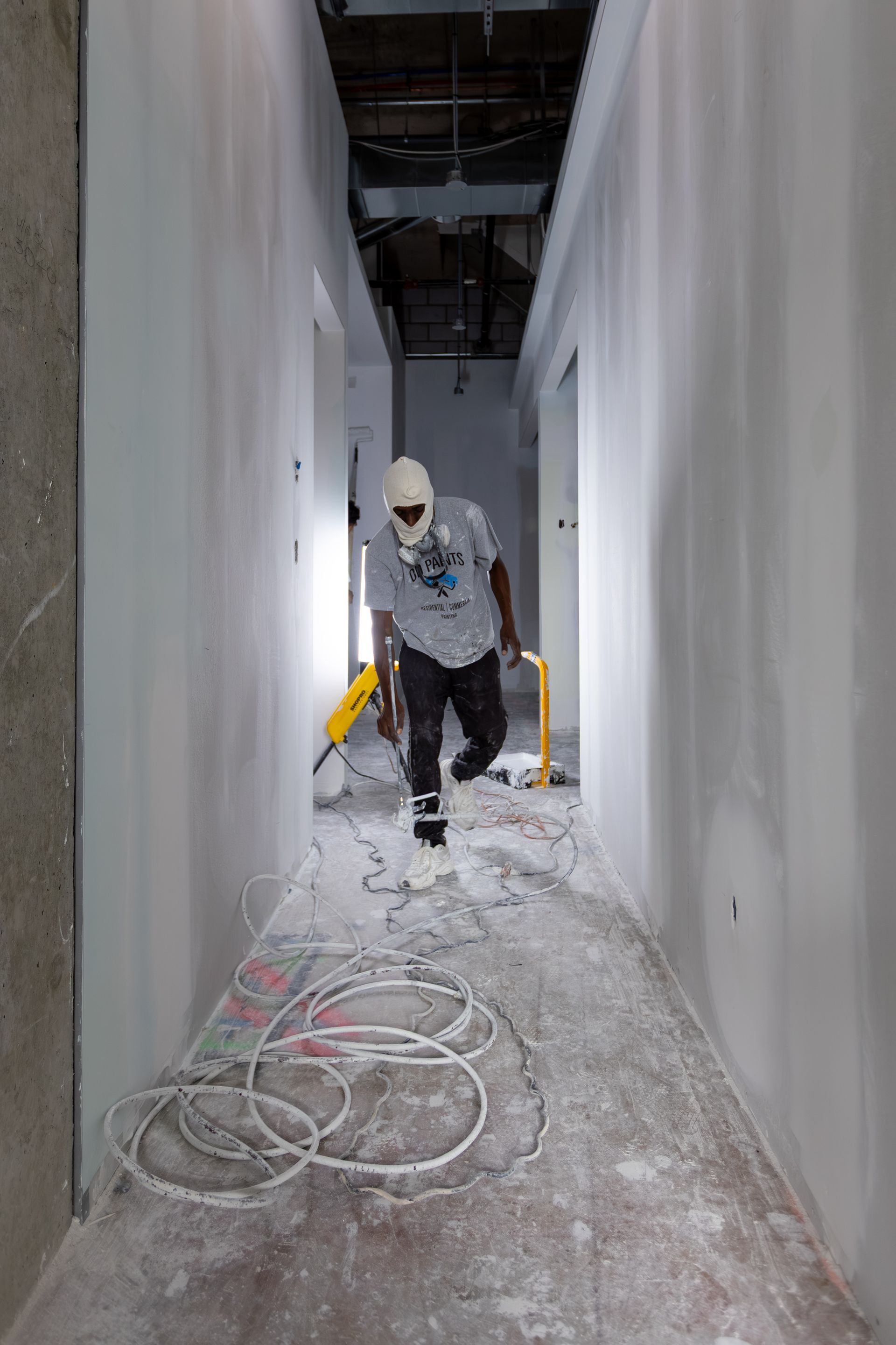 Person in a hallway during construction. Walls are white, floor is covered in dust, tools and wires are visible.
