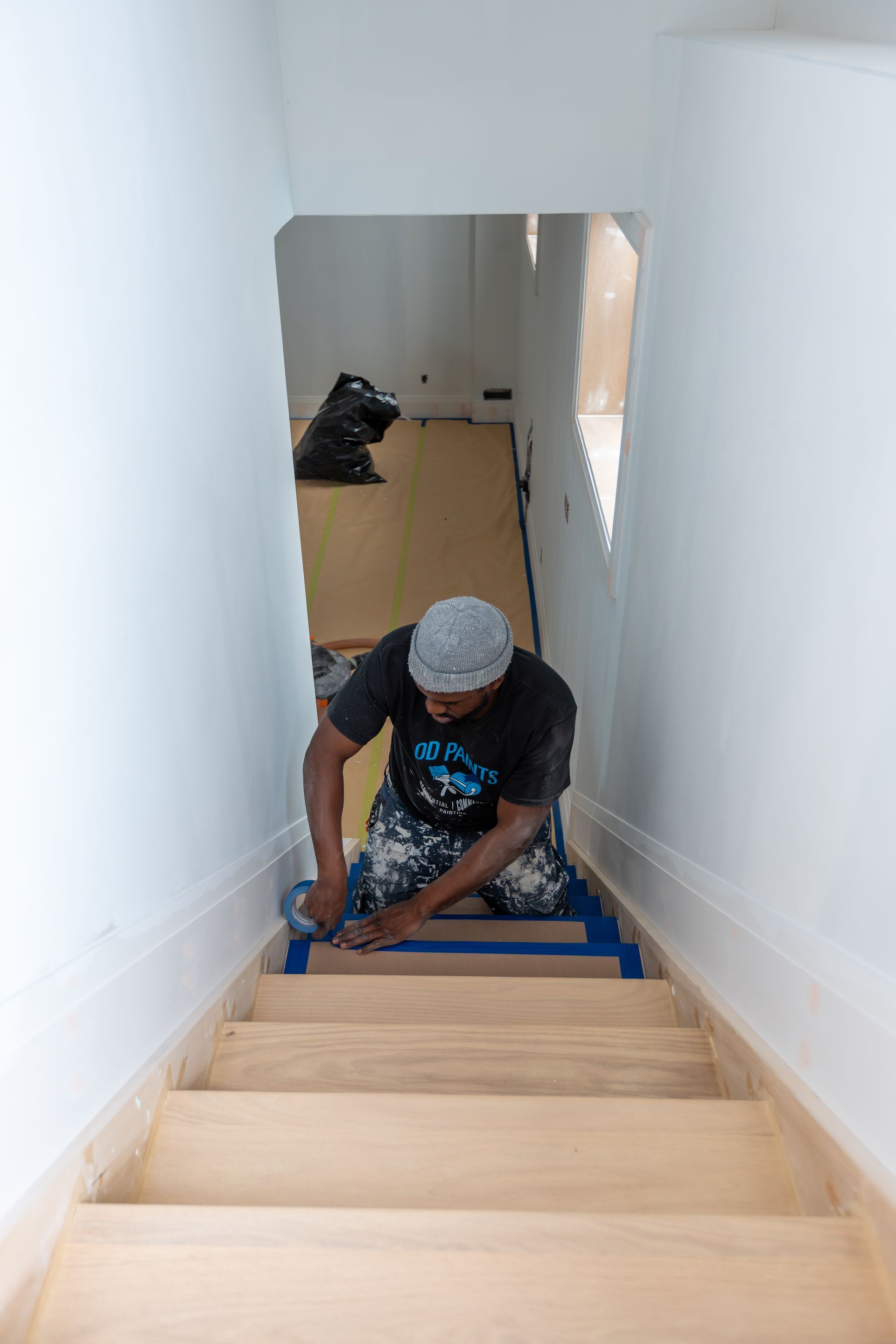 Person kneeling on staircase, taping baseboards before painting; light wood steps, white walls.