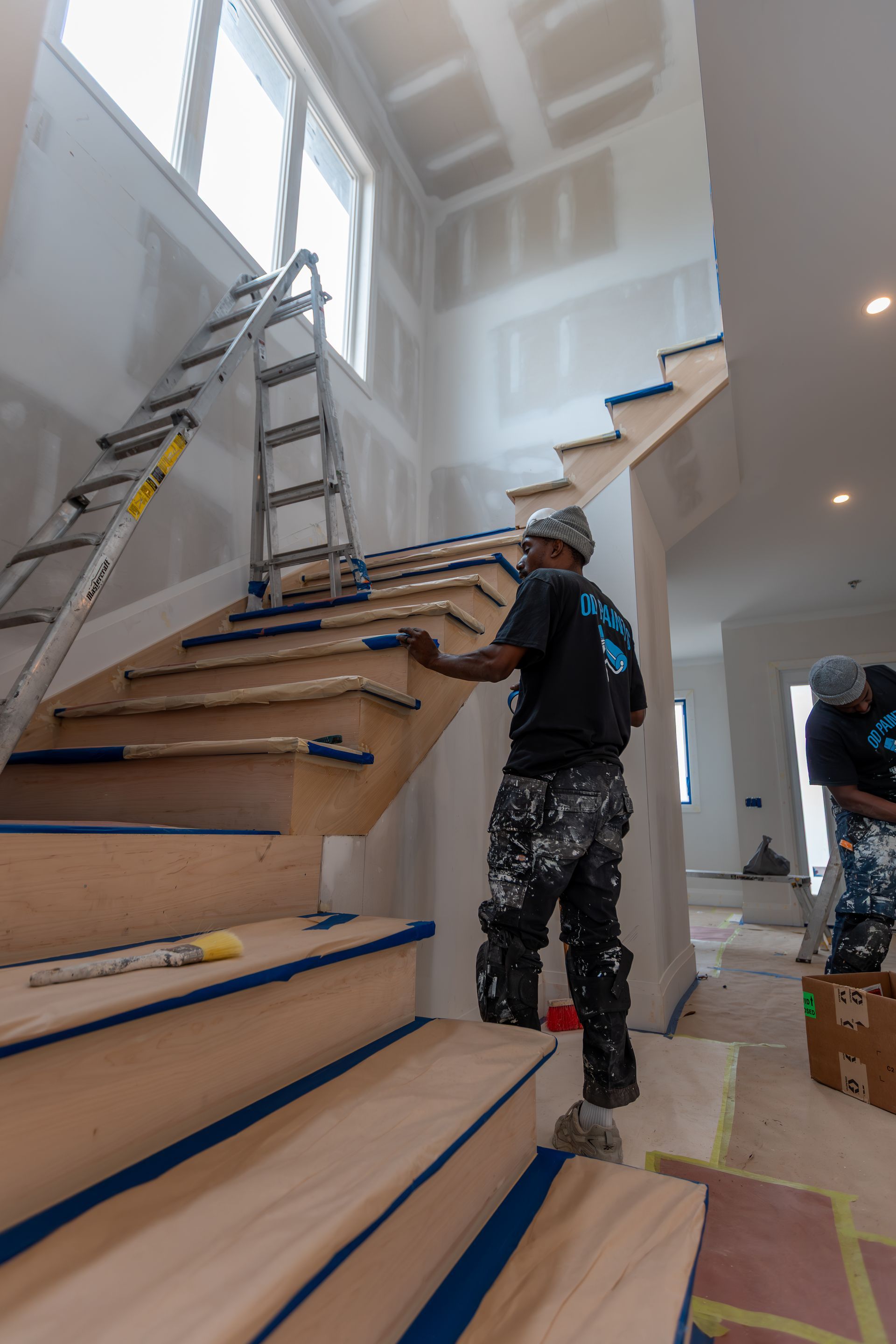 Person painting wooden staircase in a house under construction; blue tape, ladder, unfinished walls.
