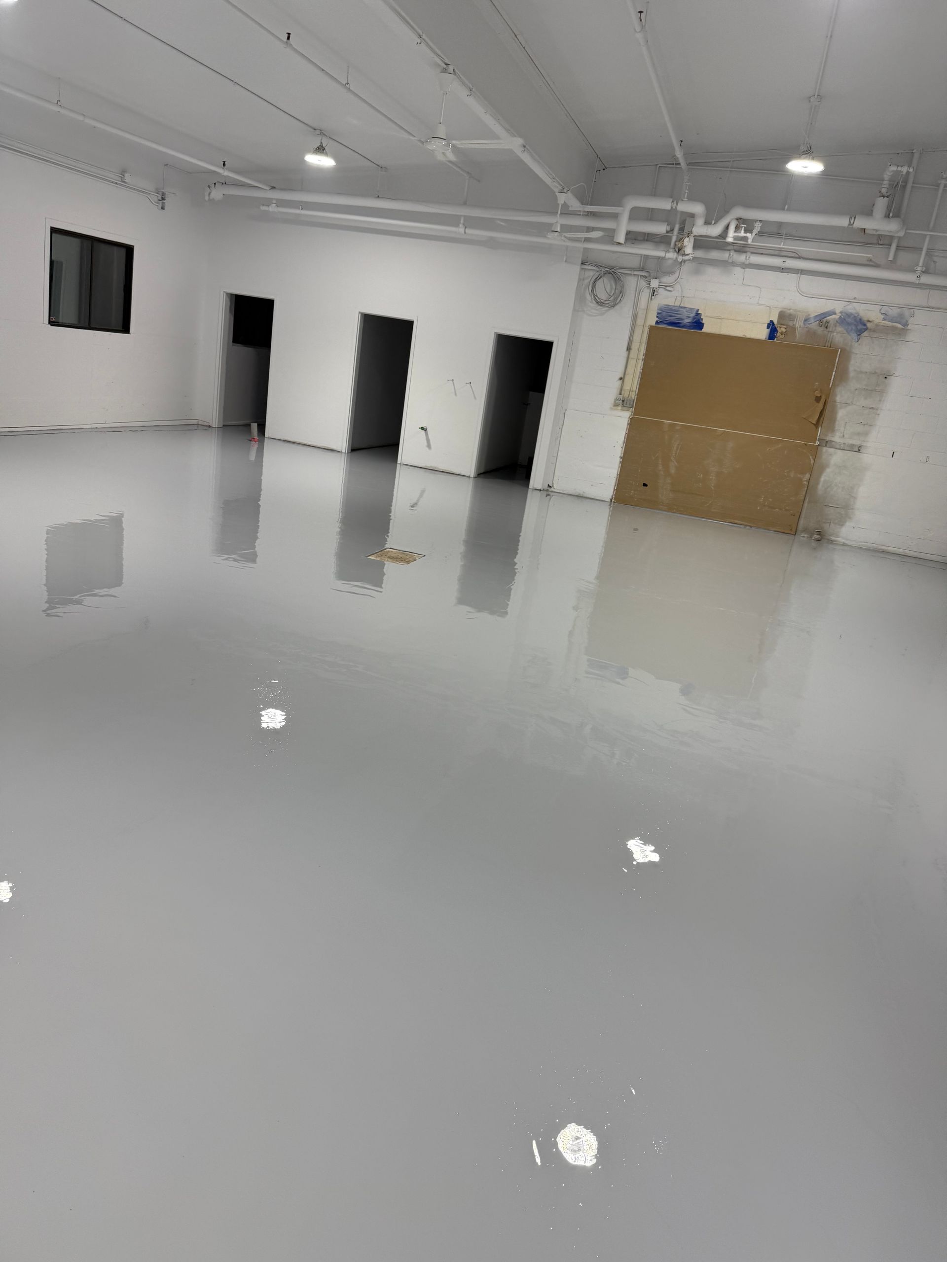 Pièce vide avec sol époxy gris brillant, trois portes et luminaires de plafond apparents.