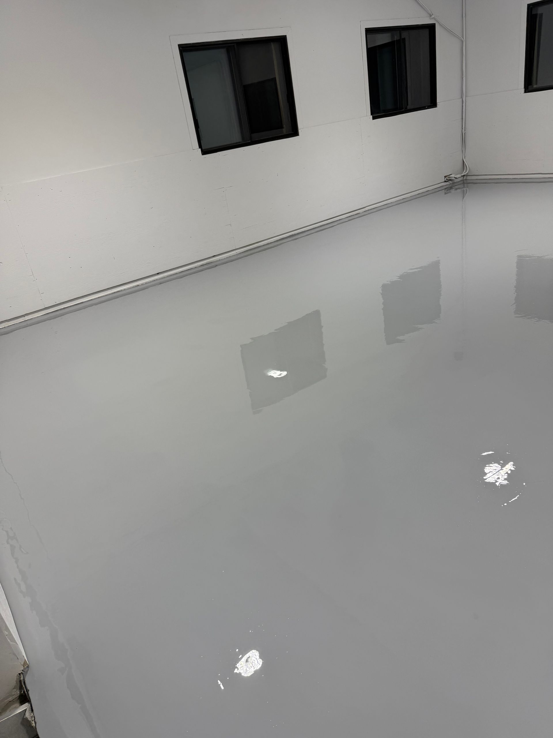 Une pièce au sol gris clair et brillant reflétant les contours carrés des fenêtres et les points lumineux. Murs blancs.