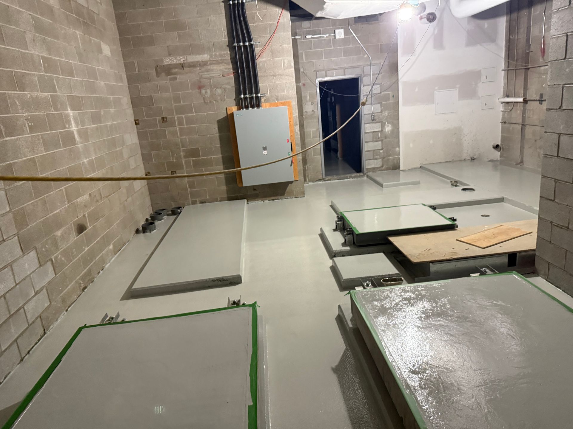 Chantier : sol en béton gris, murs en briques, porte ouverte, panneau électrique.