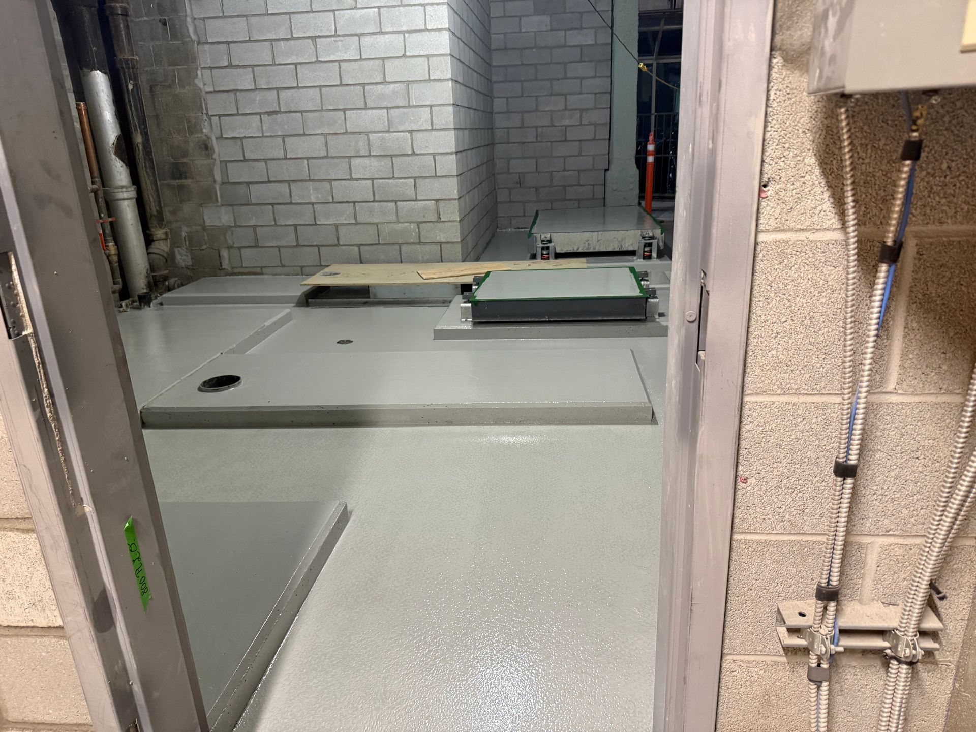 Pièce en construction au sol gris, visible depuis une porte. Murs en briques, tuyauterie et équipements sont présents.