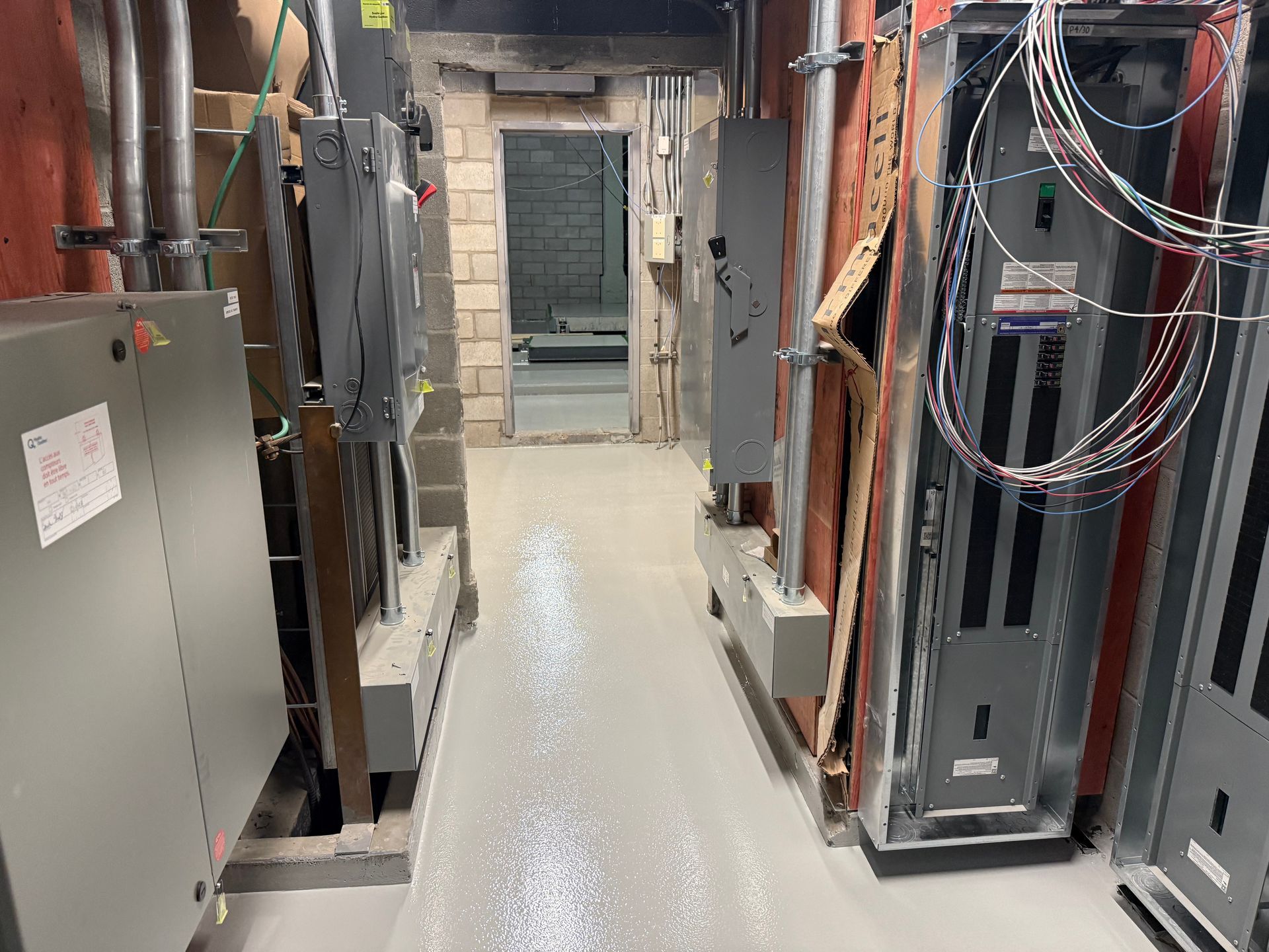 Buanderie étroite avec sol gris, boîtes métalliques, tuyaux et porte ouverte.