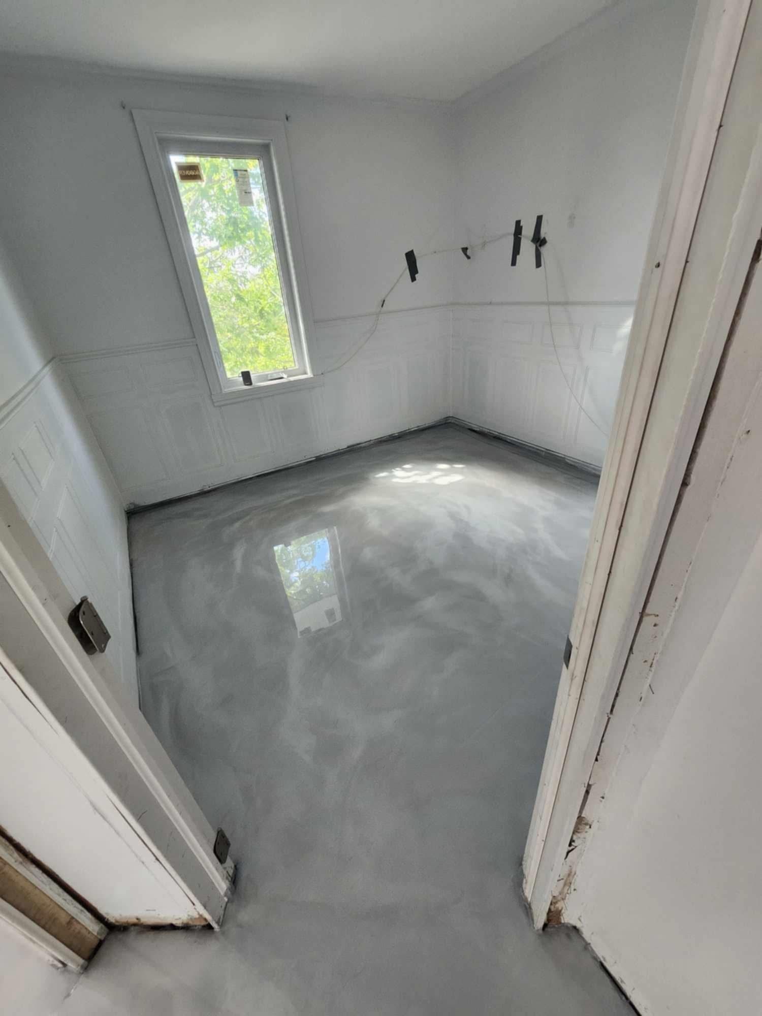 Une pièce au sol gris brillant et à la fenêtre. Les murs sont blancs et les encadrements de porte sont visibles.