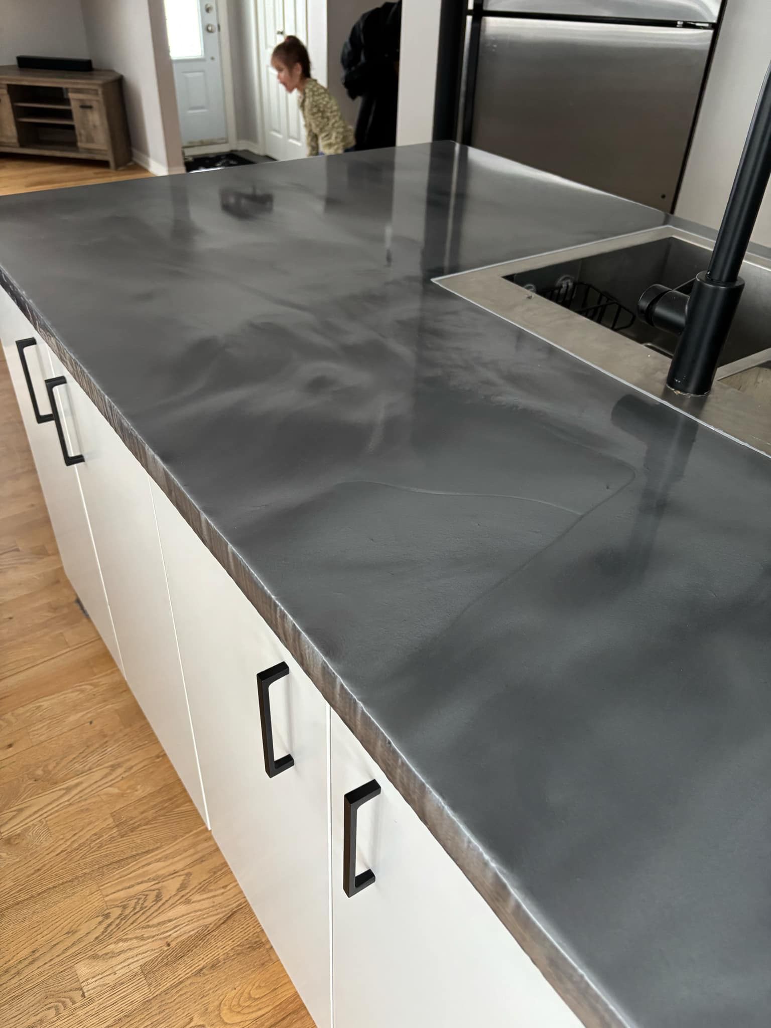 Îlot de cuisine avec plan de travail gris, armoires blanches et quincaillerie noire. Une personne jette un coup d'œil par-dessus l'îlot.