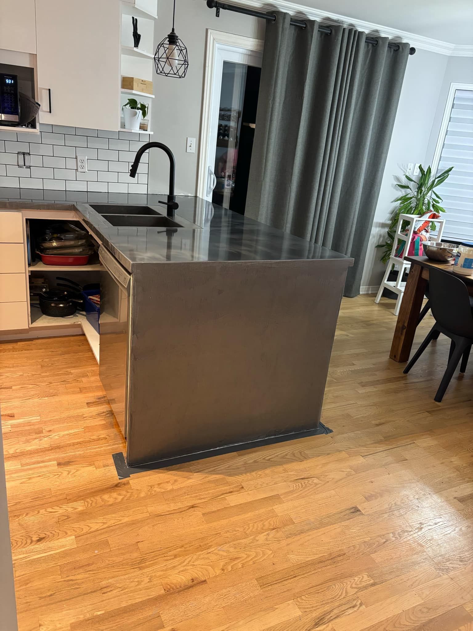 Cuisine avec îlot gris foncé, robinetterie noire et armoires blanches. Parquet, rideaux gris et lumière du jour.