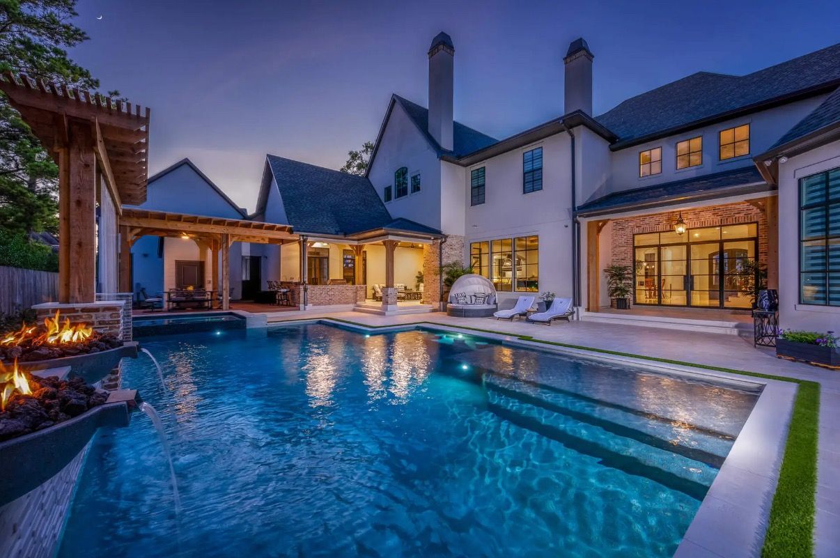Swimming pool in front of a white house on a sunny day. Lounge chairs and a shade sail are nearby.