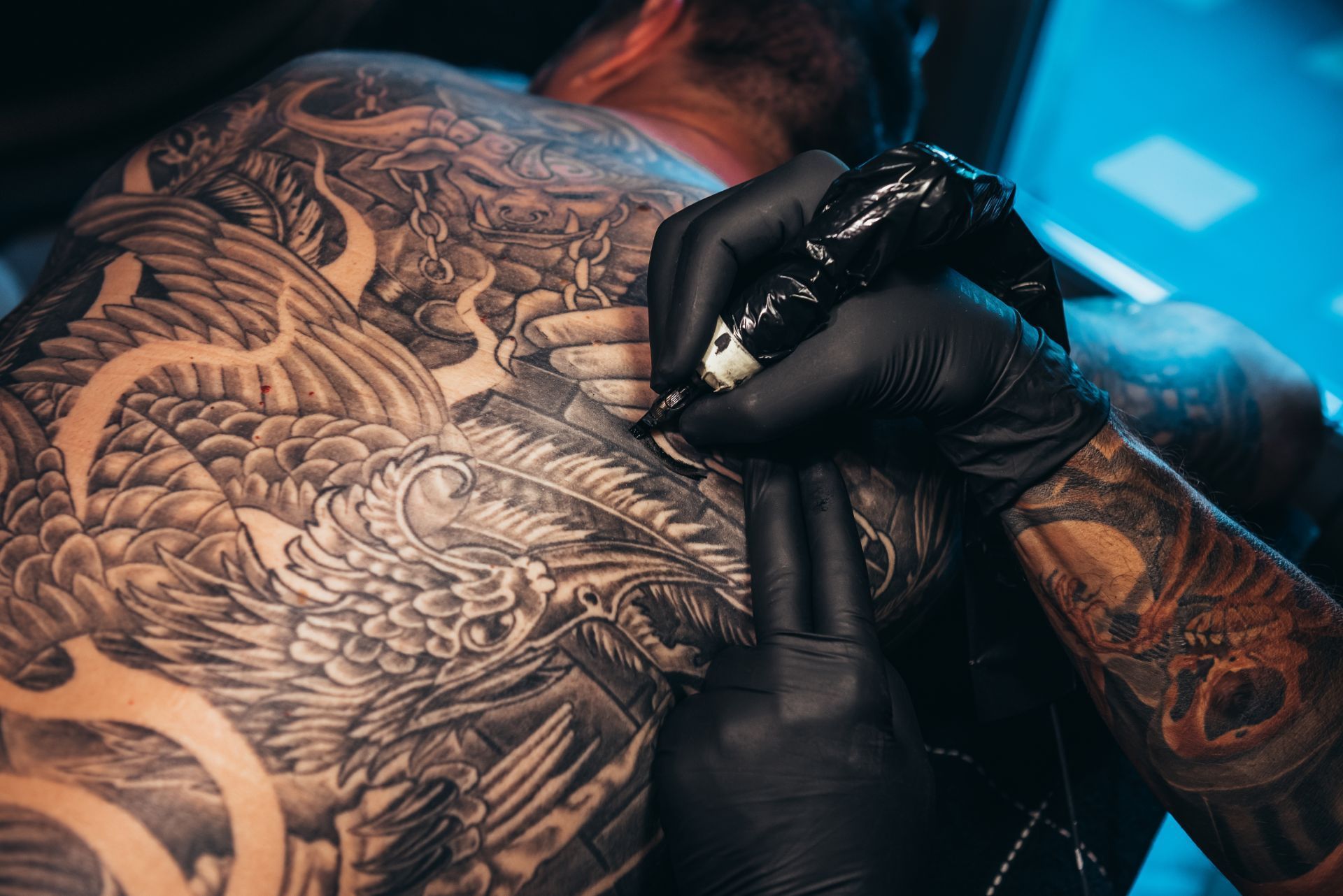 Tattoo artist in black gloves, tattooing a detailed, gray-ink design on a person's back.