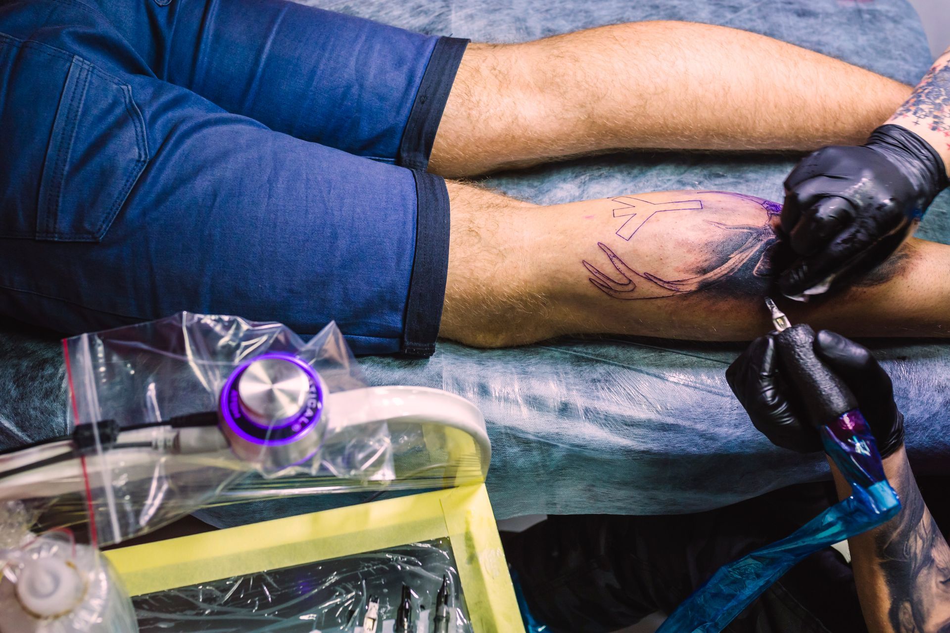 Tattoo artist with black gloves inking a leg with a tattoo machine. The leg is blue-jeaned.