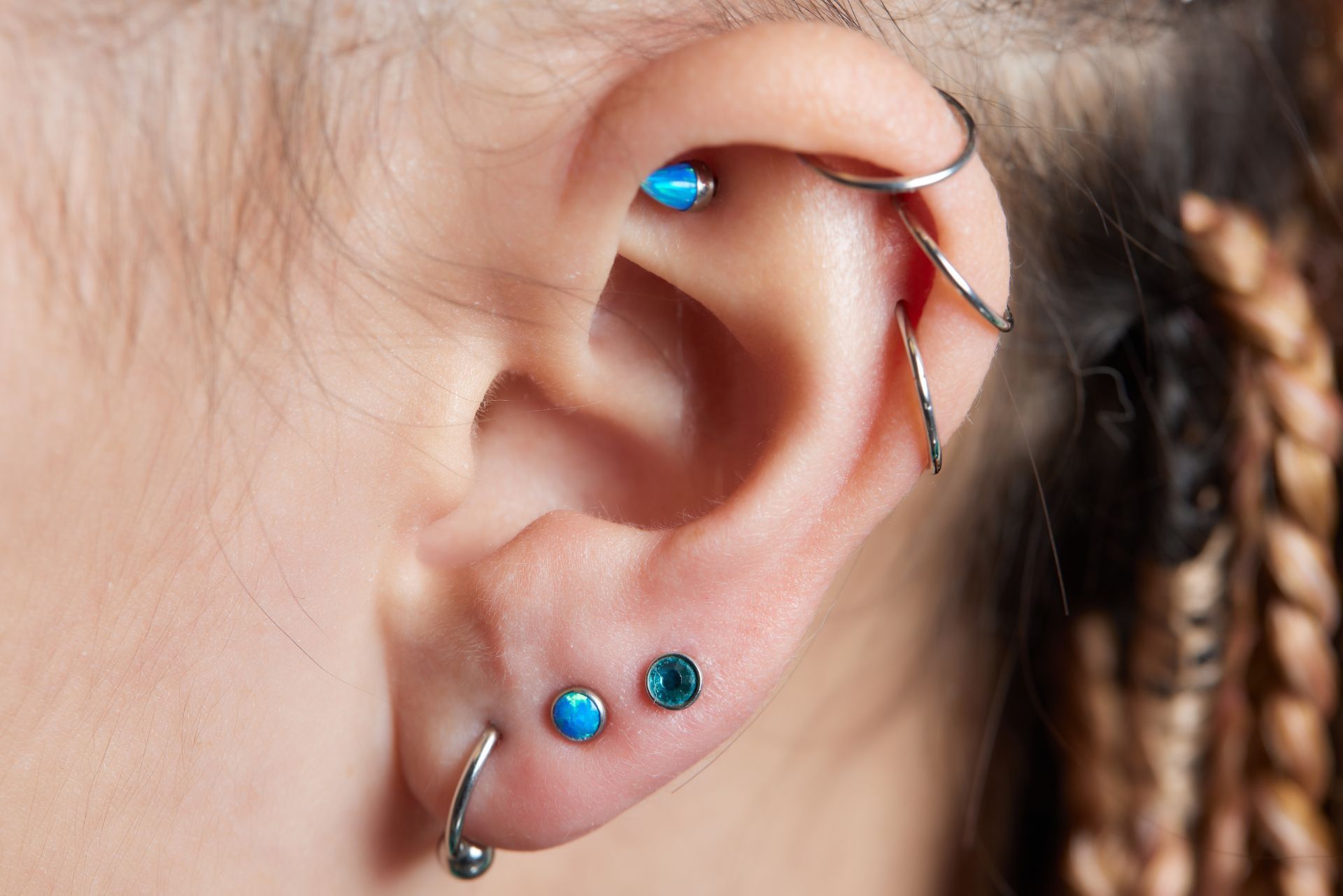 Ear with multiple piercings: helix, tragus, and lobe with silver and blue-gem jewelry. Braids visible.