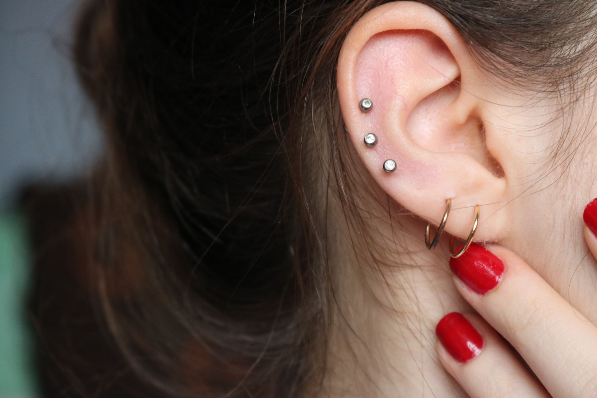 Ear with multiple piercings: helix studs and gold hoop earrings, red nails.