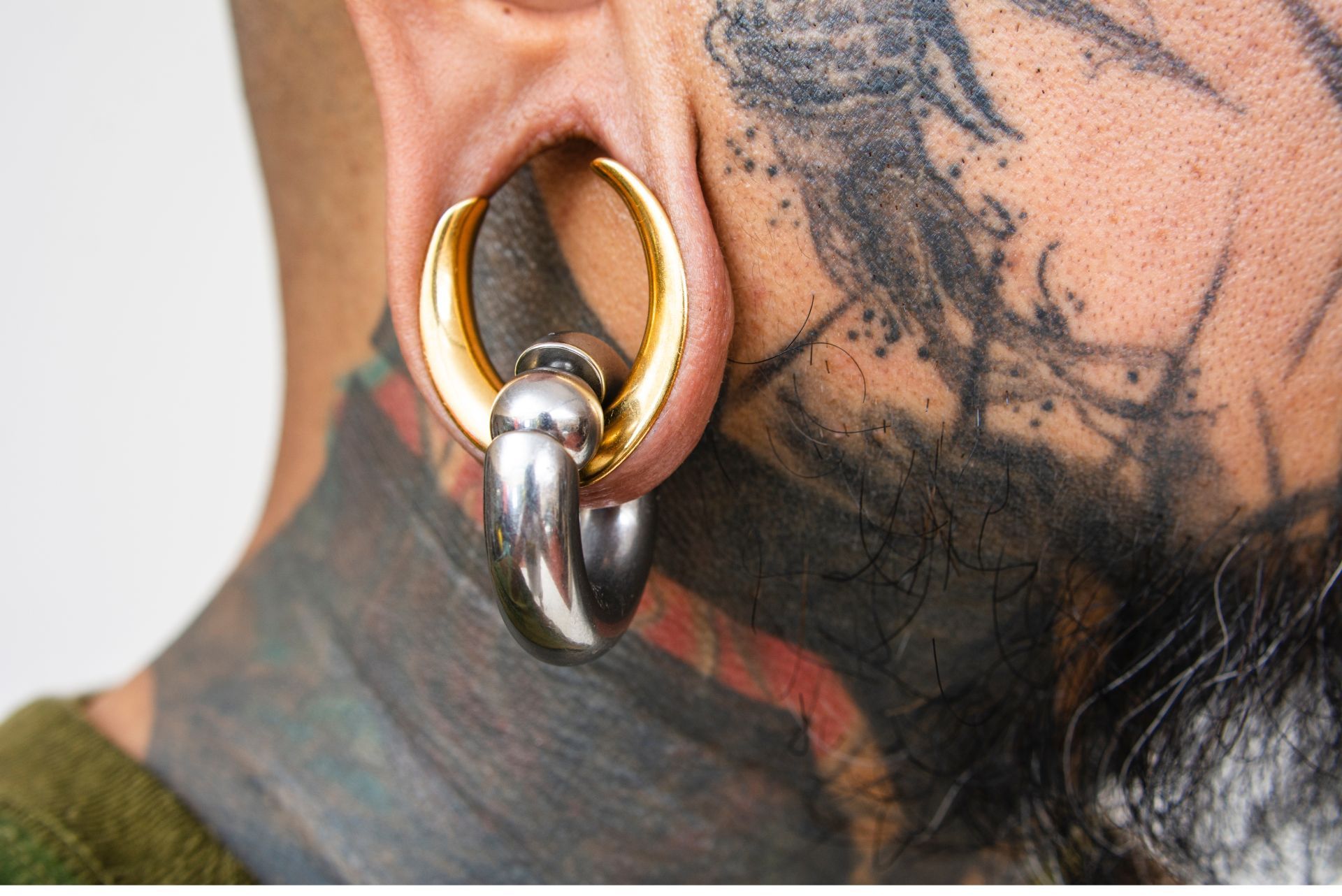 Close-up of a person's ear with a large, golden and silver ear gauge. Visible neck and face tattoos.