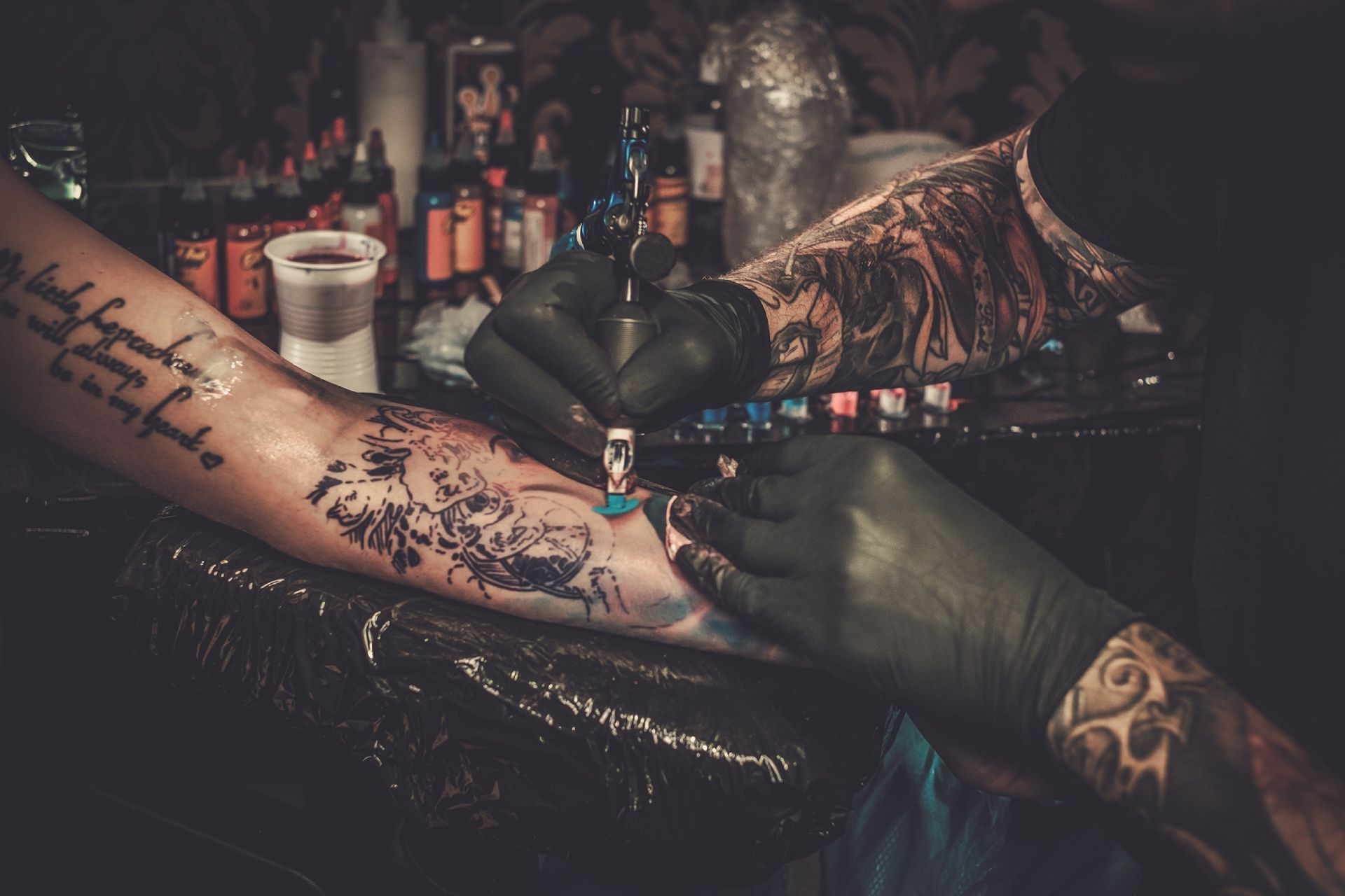 Tattoo artist with black-gloved hands, tattooing a detailed design on a person's forearm.