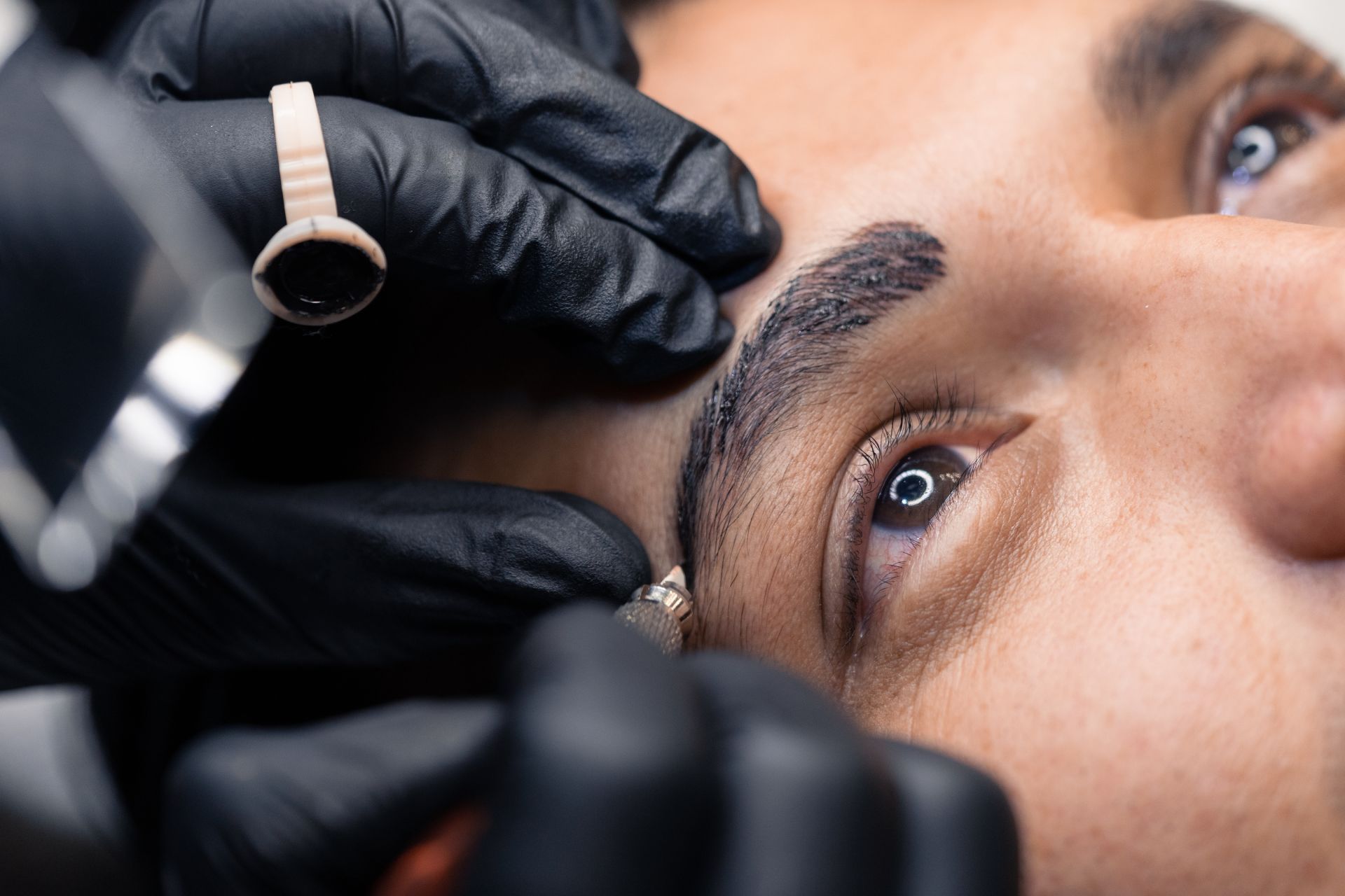 Person receiving eyebrow microblading. Black gloved hands holding tools near eye. Brown eyebrow partially shaded.