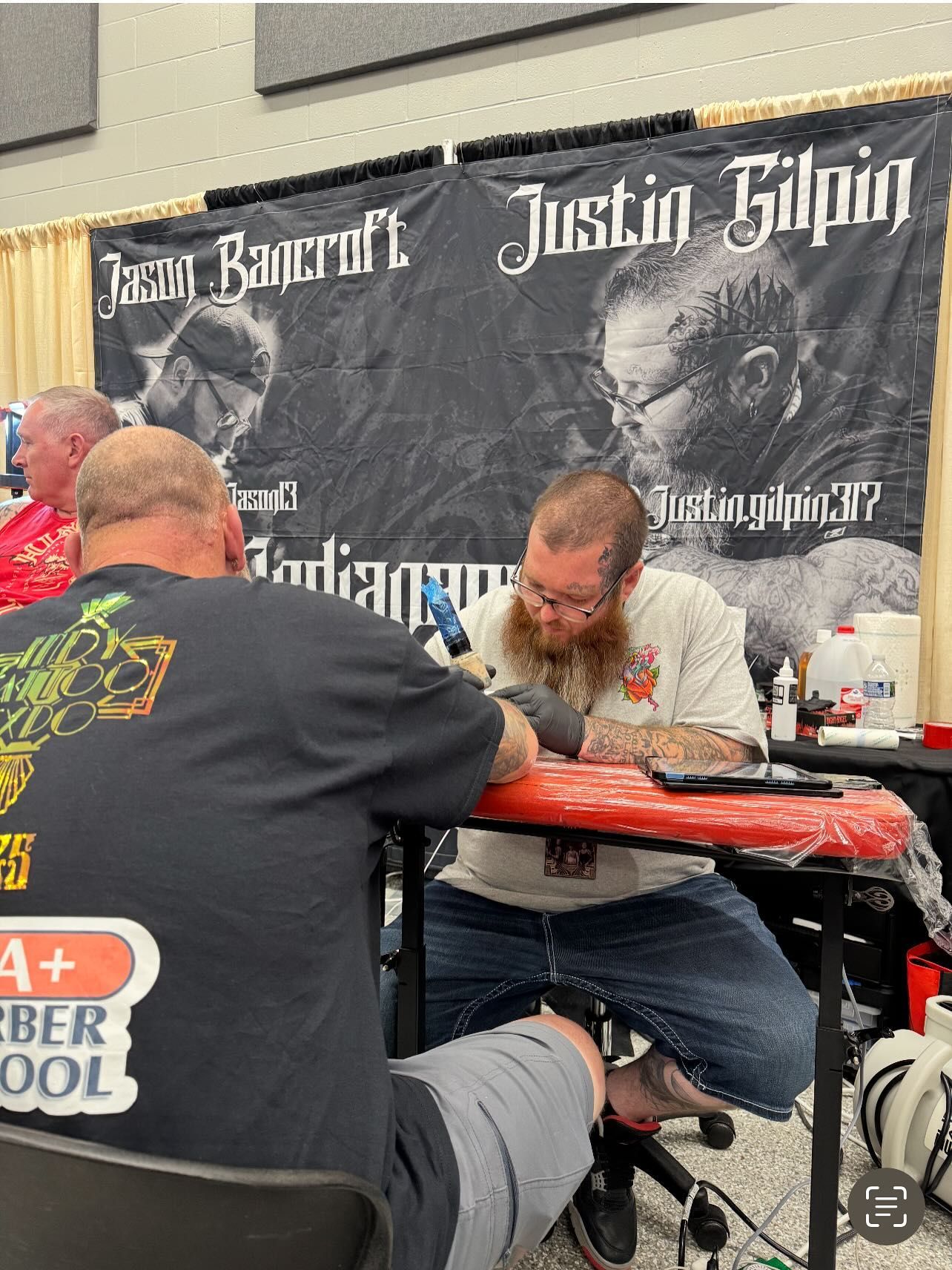 Tattoo artist working on a client at a convention; banner with names.