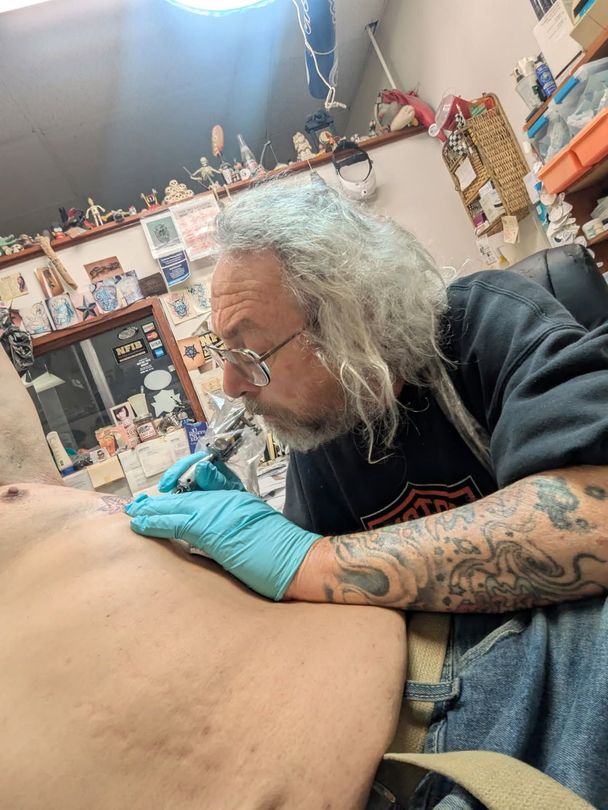 Tattoo artist with gray hair and glasses tattoos torso in cluttered shop.
