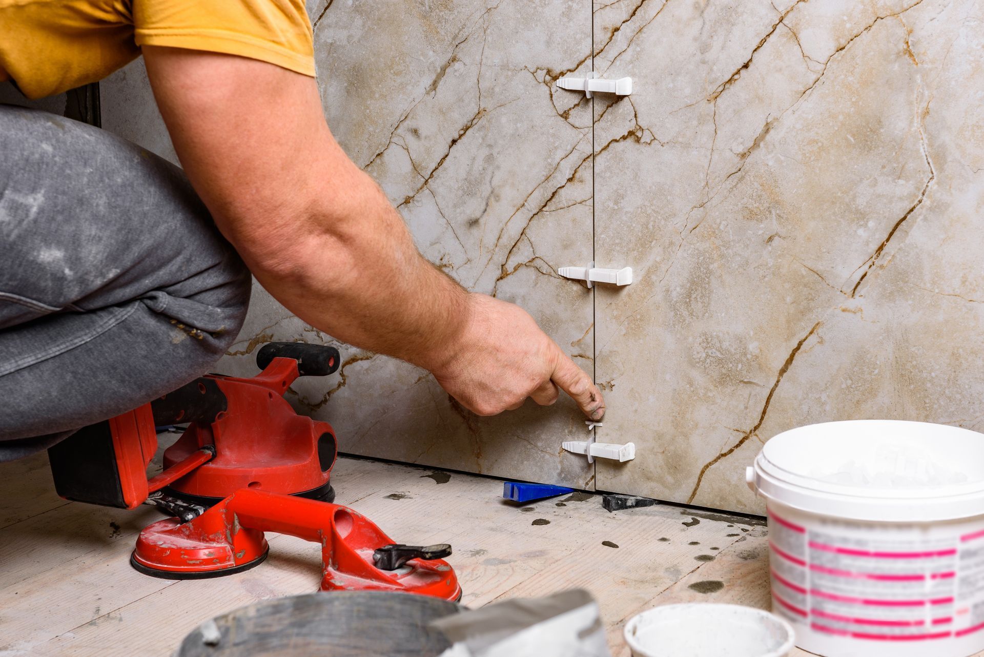 A man is kneeling down while installing tiles on a wall.