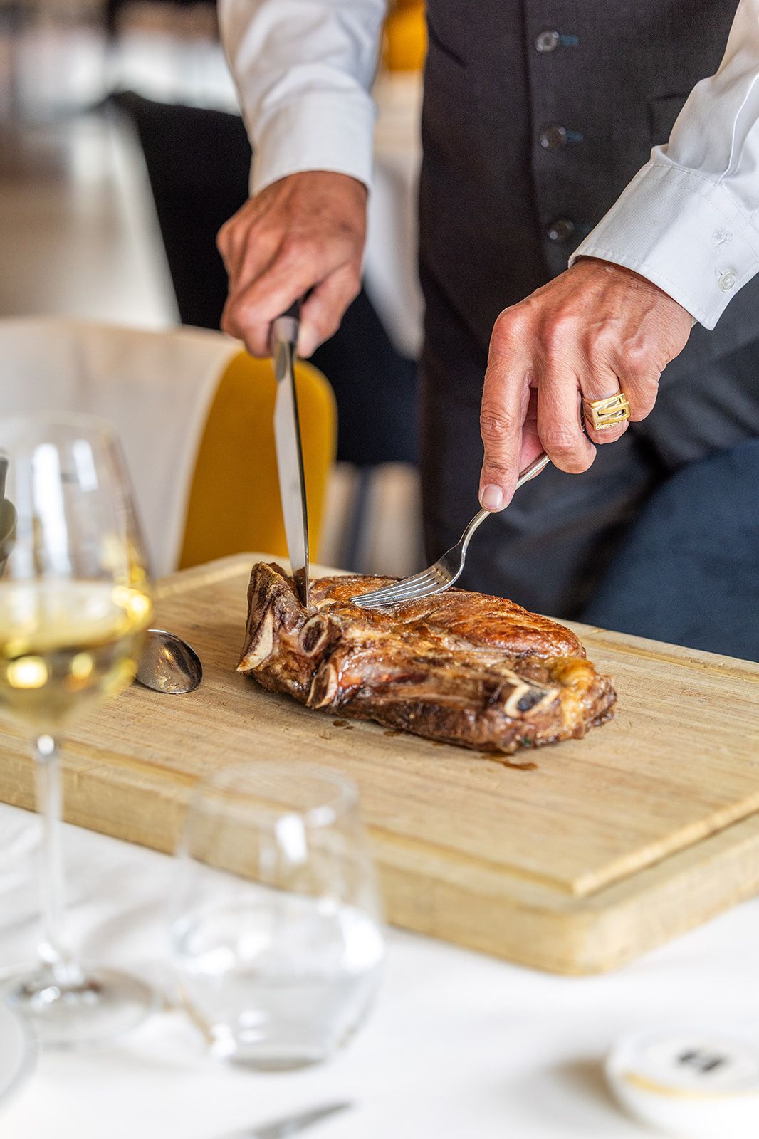 A chef in a dark apron and cap smiles while plating a green sauce from a small pan in a professional kitchen.