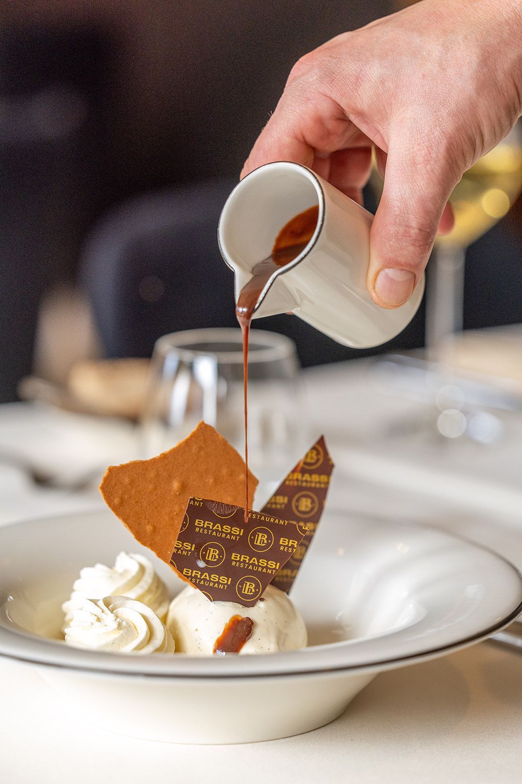 Hand pouring sauce over a plated dessert with whipped cream and chocolate garnish
