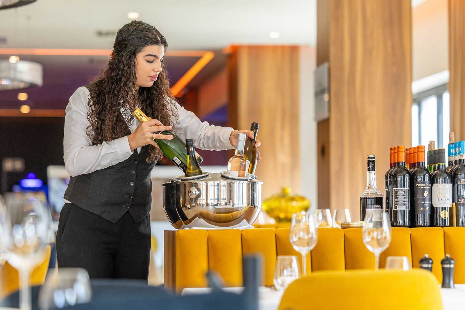 A bright, modern restaurant with wood floors, exposed brick, large windows, and yellow upholstered chairs at tables.