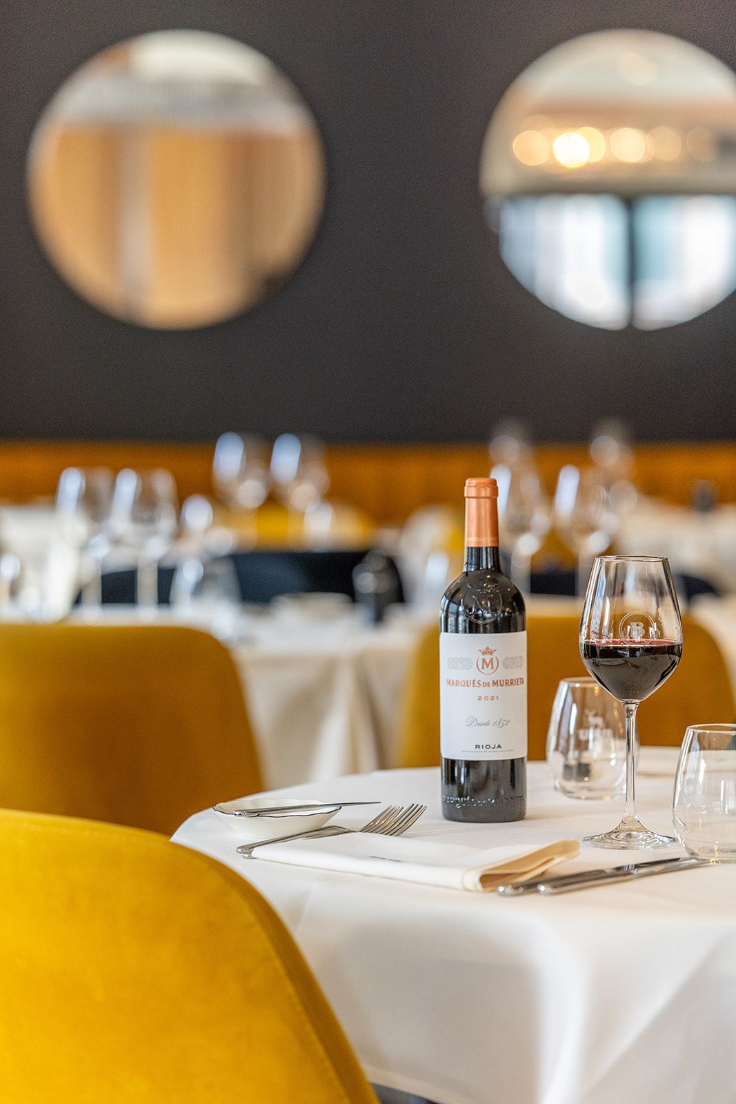 Three smiling staff members in patterned blouses or a shirt and grey aprons stand together inside a brick-walled space.