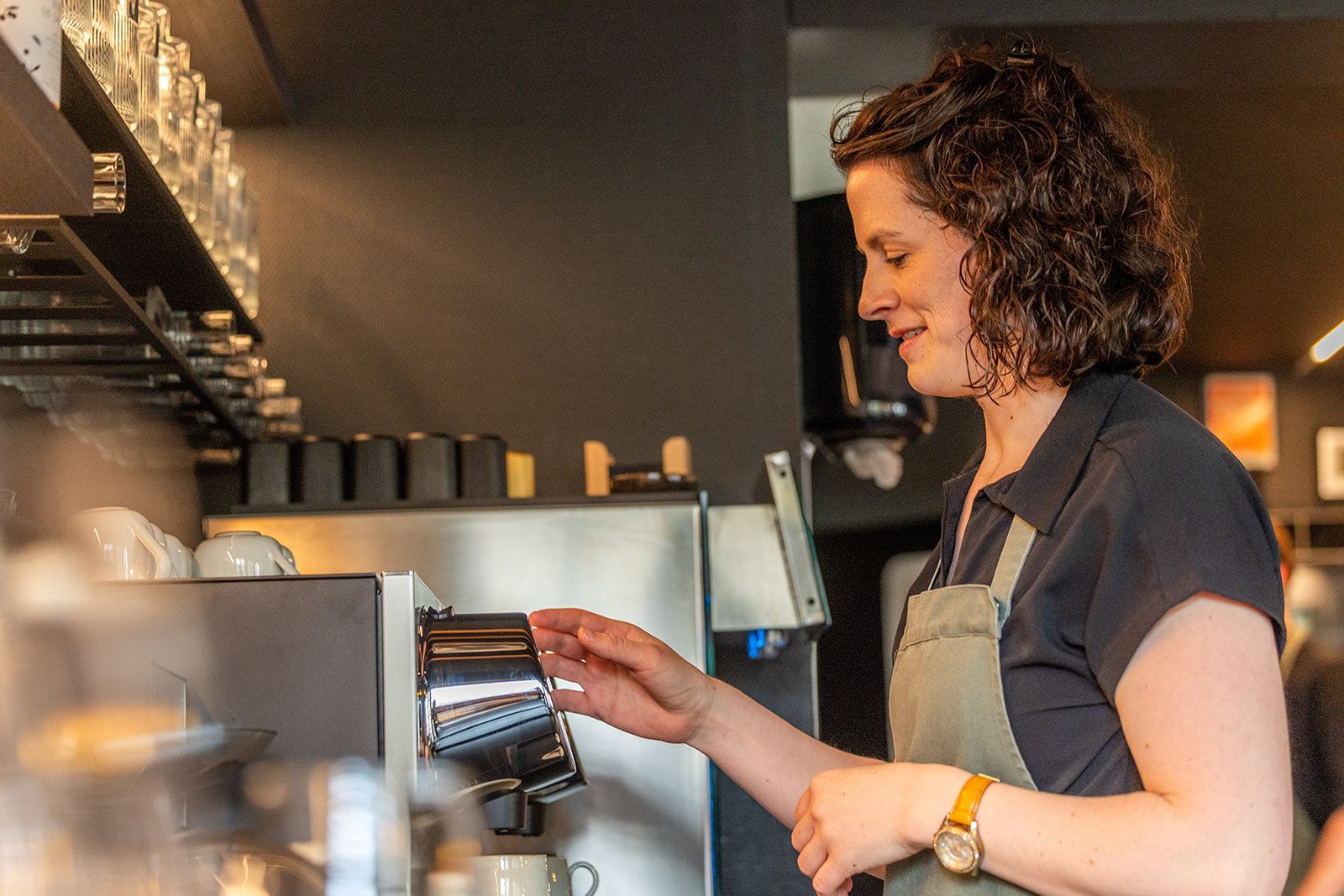 A smiling person stands in a restaurant wearing a patterned blouse and apron, holding two small plates of food.