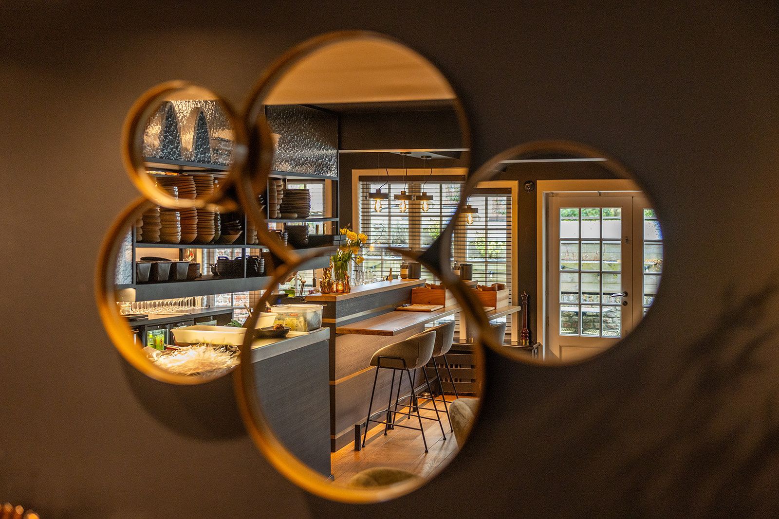 A chef in a dark apron and cap smiles while plating a green sauce from a small pan in a professional kitchen.