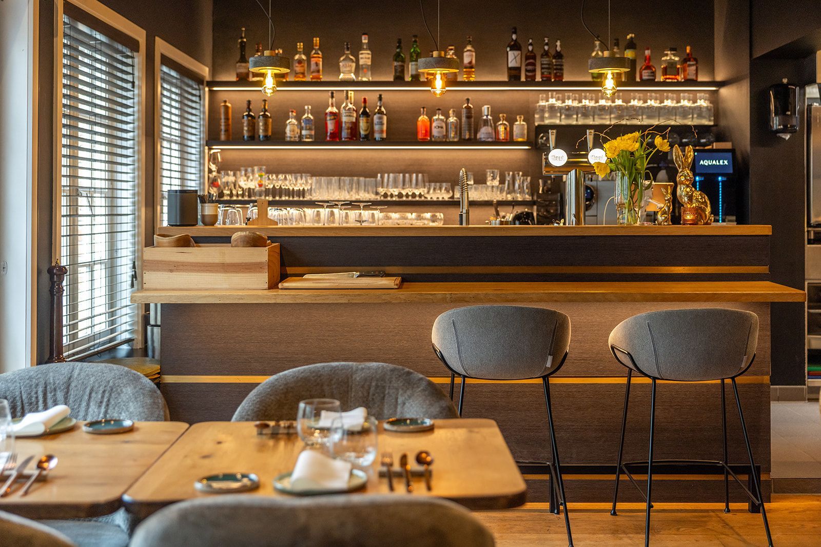 A bowl of green pesto pasta next to a wooden paddle with small appetizers on a leather placemat with glasses.