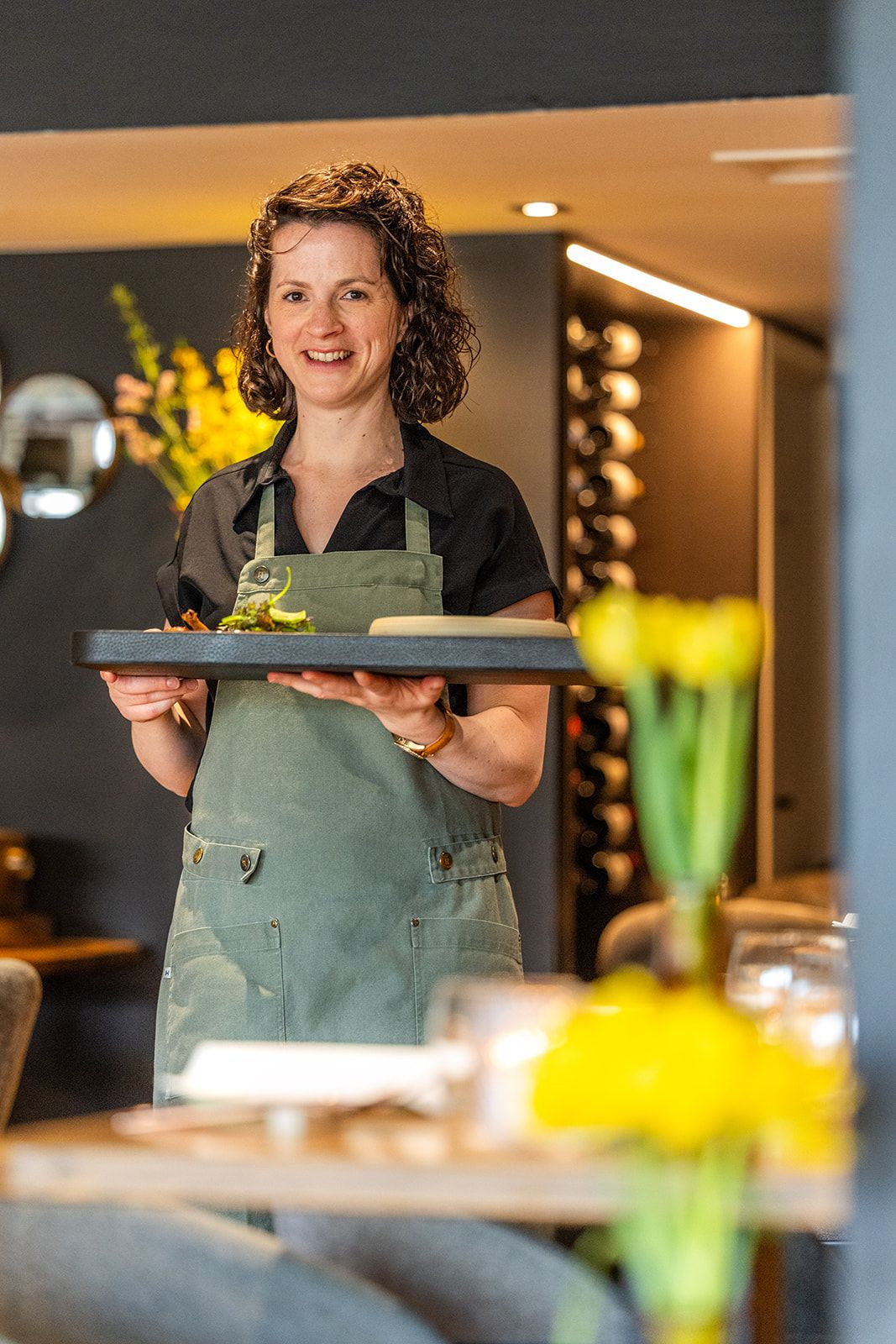 A table set for dining in a restaurant with exposed brick walls, a window overlooking a bar, and yellow and green chairs.