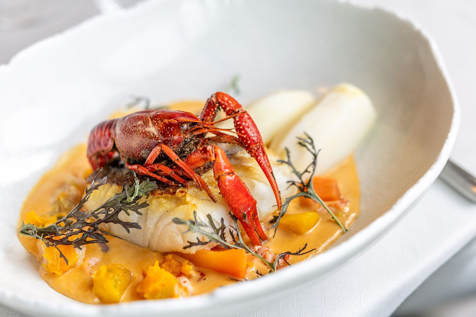 A chef in a dark apron and cap smiles while plating a green sauce from a small pan in a professional kitchen.