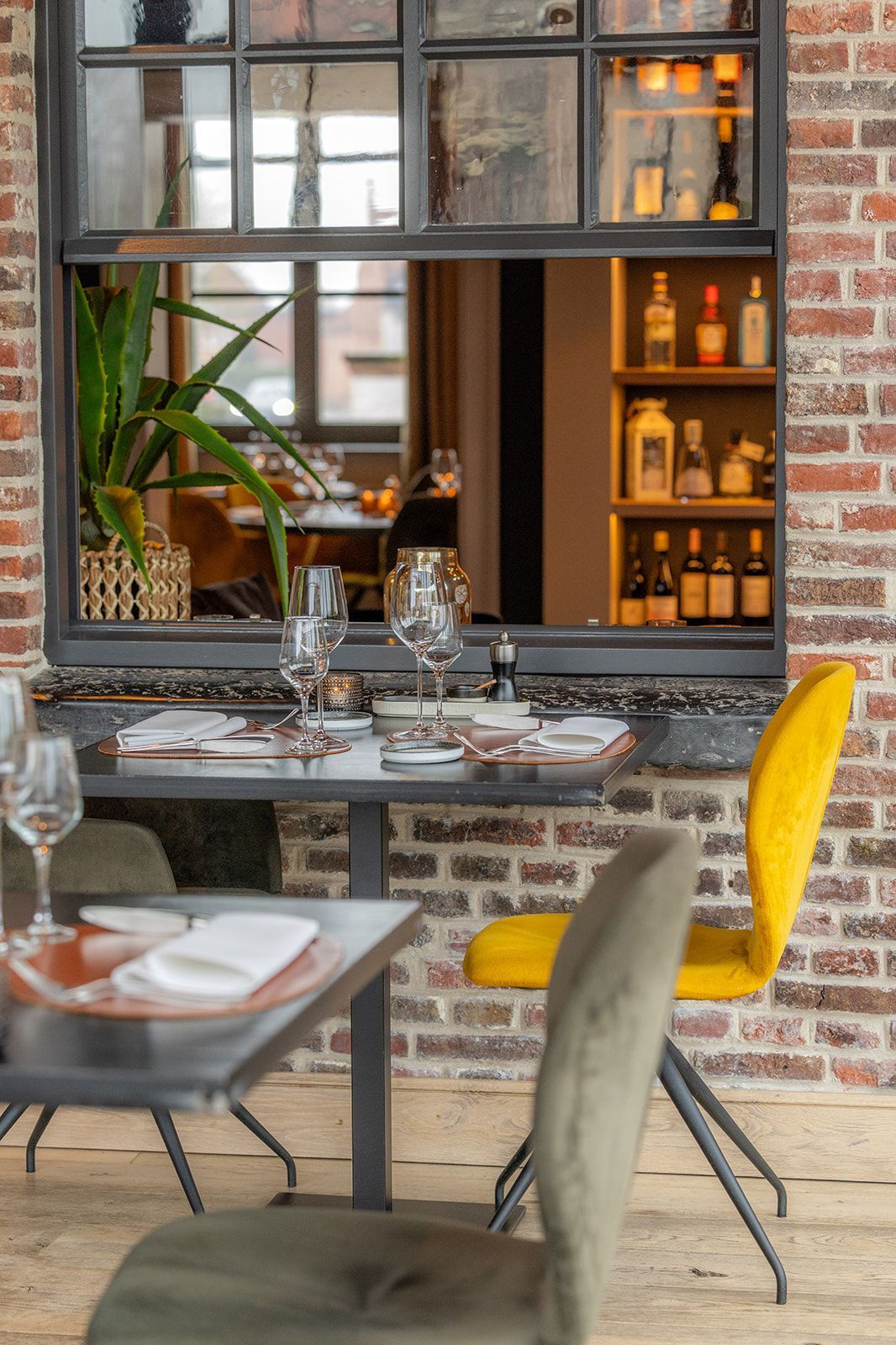 A table set for dining in a restaurant with exposed brick walls, a window overlooking a bar, and yellow and green chairs.