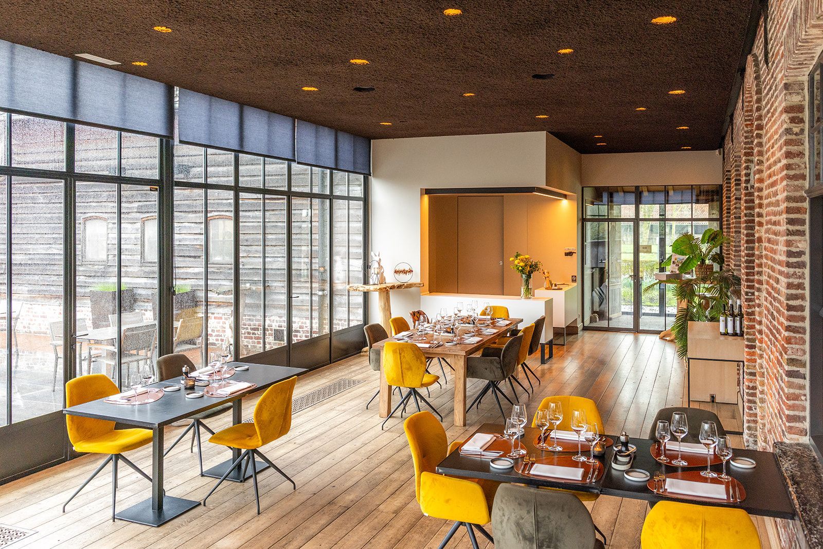 A dining room with round tables set with white tablecloths, folded napkins, and plates, with large windows in the back.