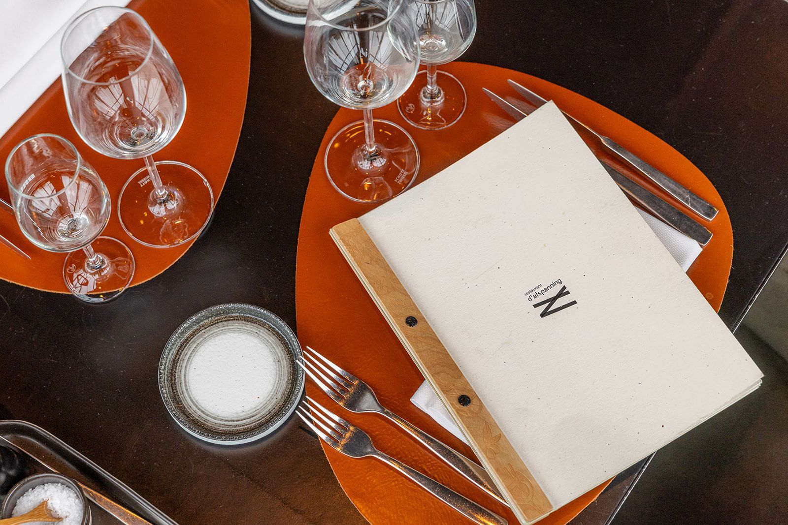A white menu with a wooden spine rests on a vibrant orange placemat, accompanied by wine glasses and cutlery on a table.