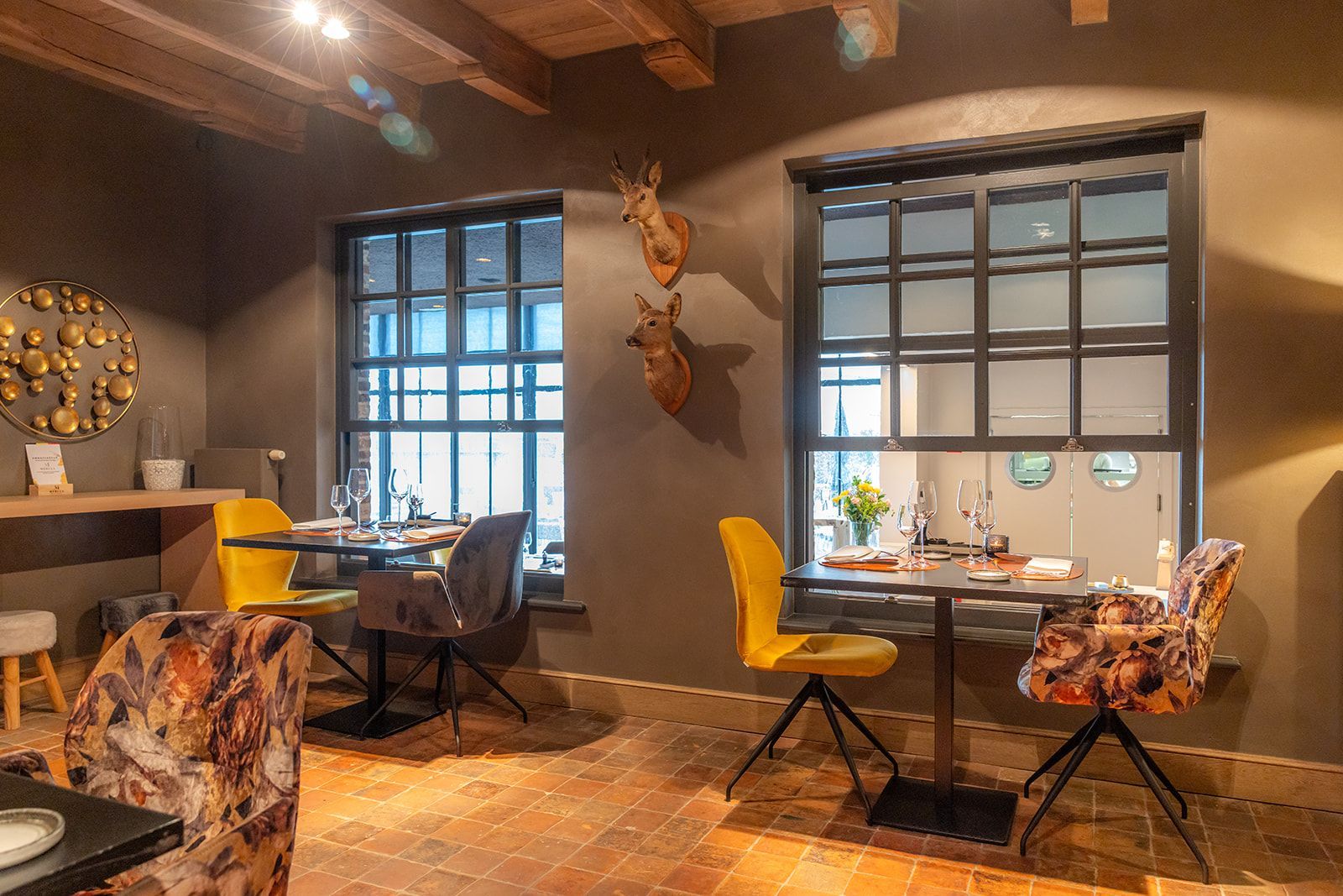 A restaurant interior with two tables, yellow and patterned chairs, brick flooring, and deer head wall decor.