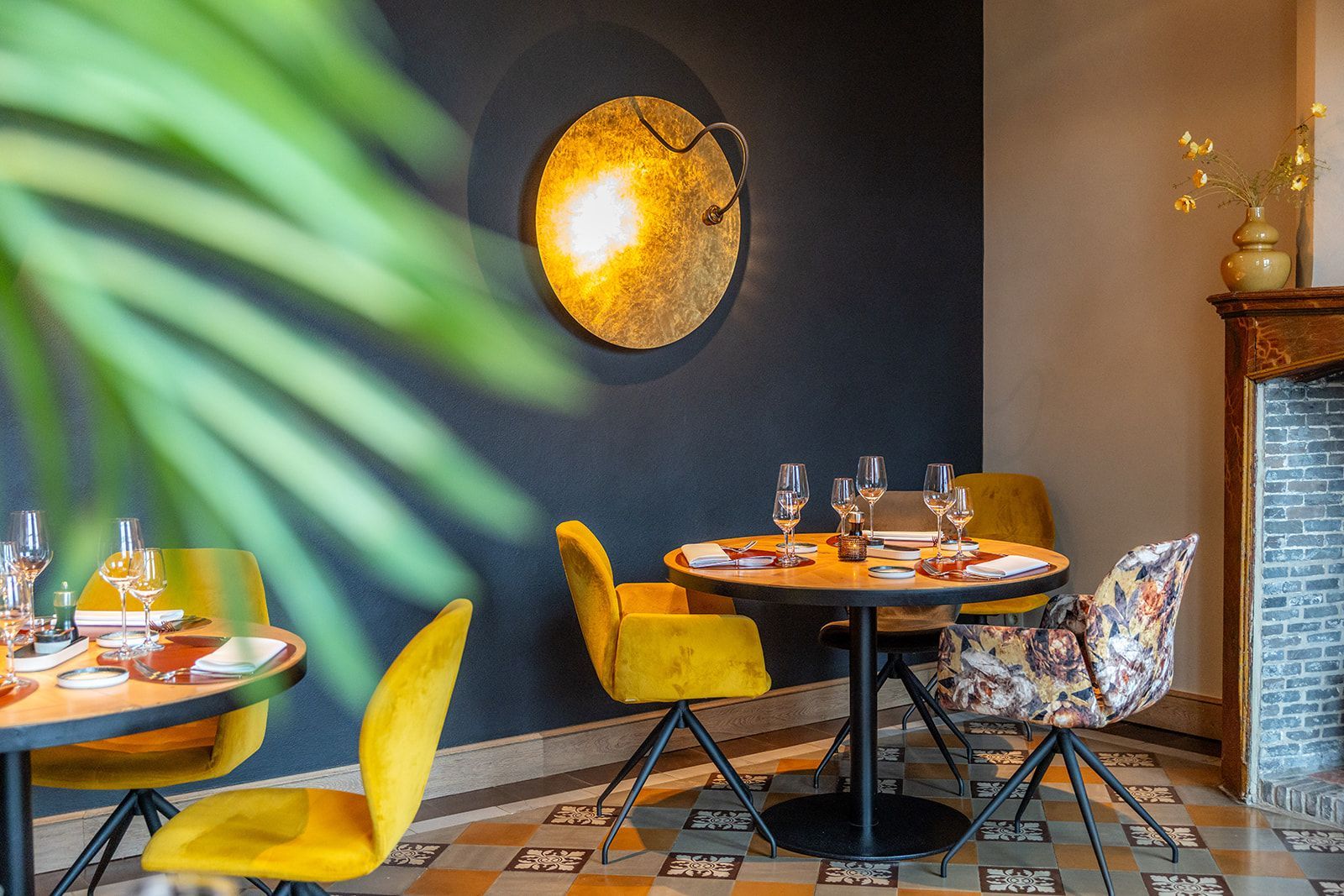 A restaurant interior featuring round tables with yellow chairs, a dark blue wall with a round gold light, and patterned tiles.