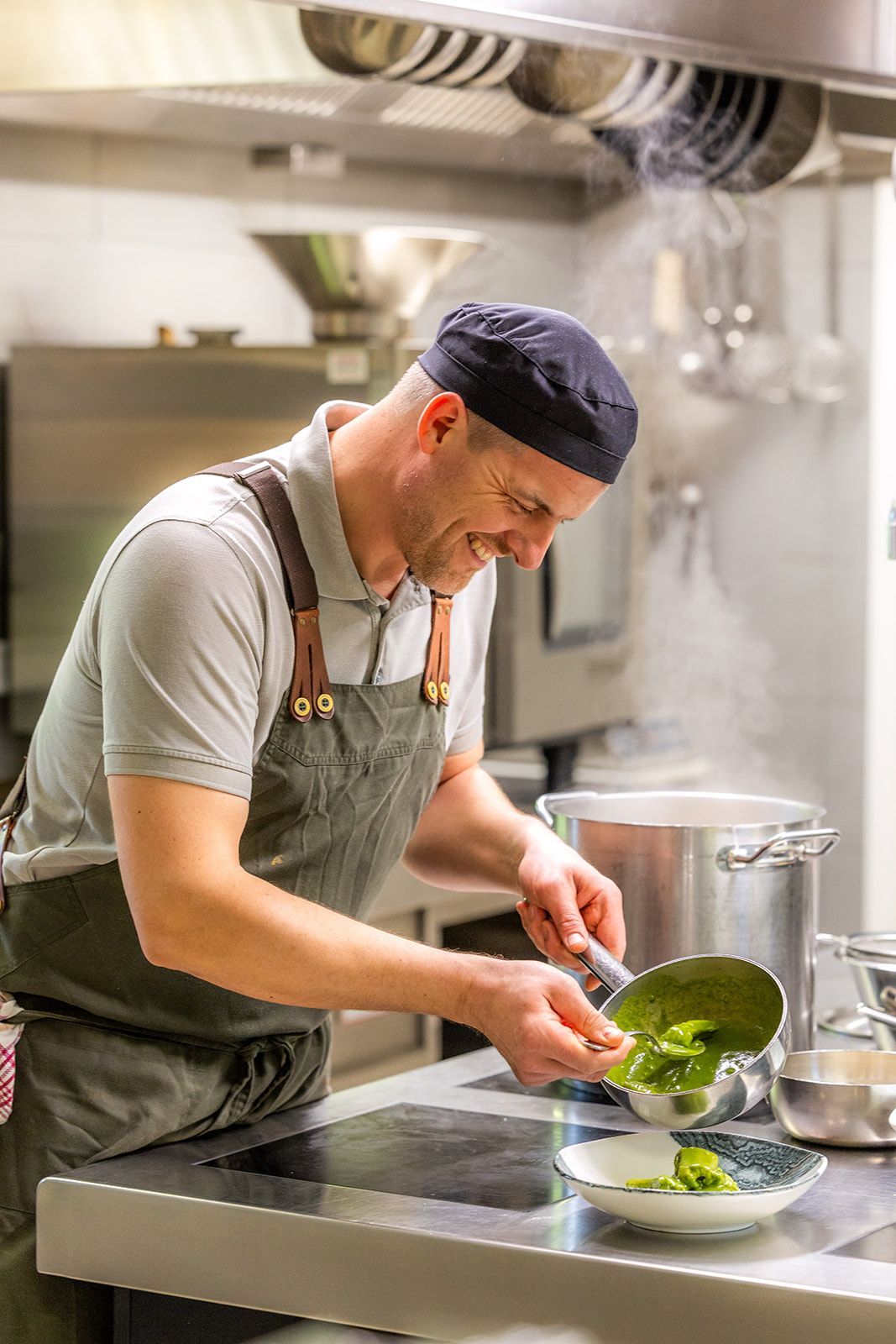 A chef in a dark apron and cap smiles while plating a green sauce from a small pan in a professional kitchen.